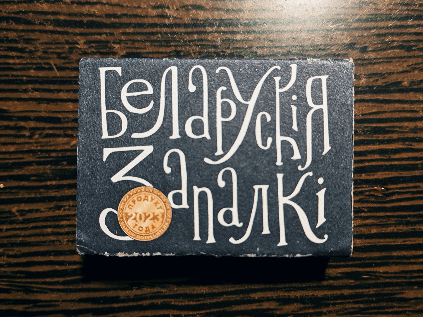 Коробок с белорусскими спичками, на темном фоне белый текст: «Беларускі Запалкі» и круглая печать.