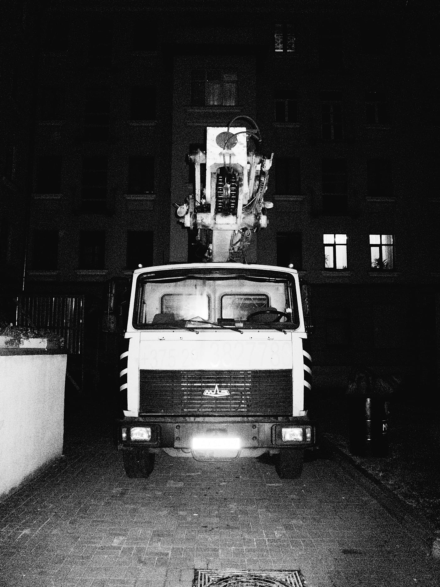 Автокран стоит в ночном дворе возле дома. Чёрно белая фотография. Снято со вспышкой.
