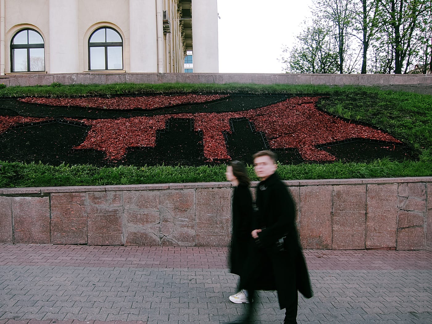 Парень и девушка в чёрном проходят на фоне весенней клумбы в центре Минска