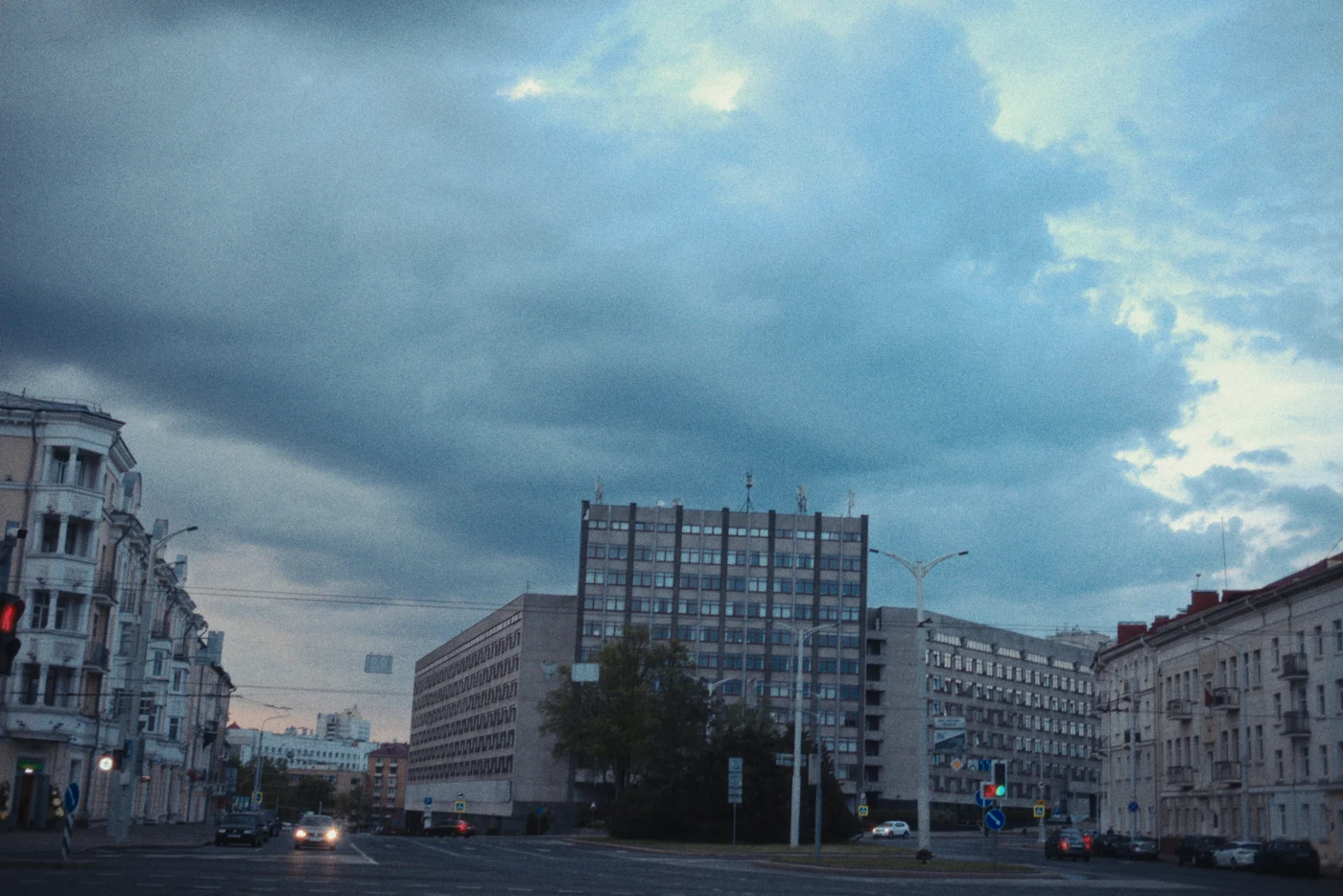 Городская улица с серыми облаками, зданиями и светофором. На горизонте виднеется современный многоэтажный дом.