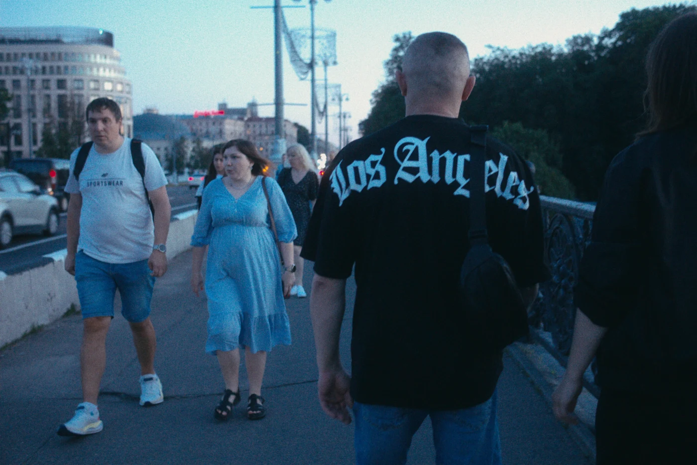 Группа людей на улице, один в футболке с надписью «Los Angeles», вечернее освещение.