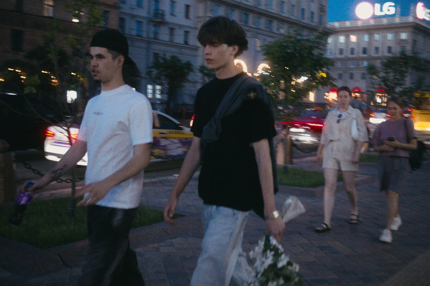 Молодые люди идут по улице, один с букетом цветов, вечерняя атмосфера города.