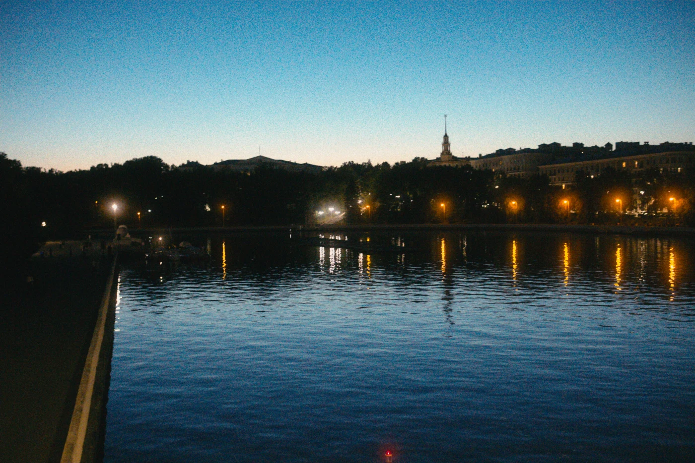 Ночной пейзаж с рекой и отражением огней на воде, на фоне silhouetted зданий и неба.