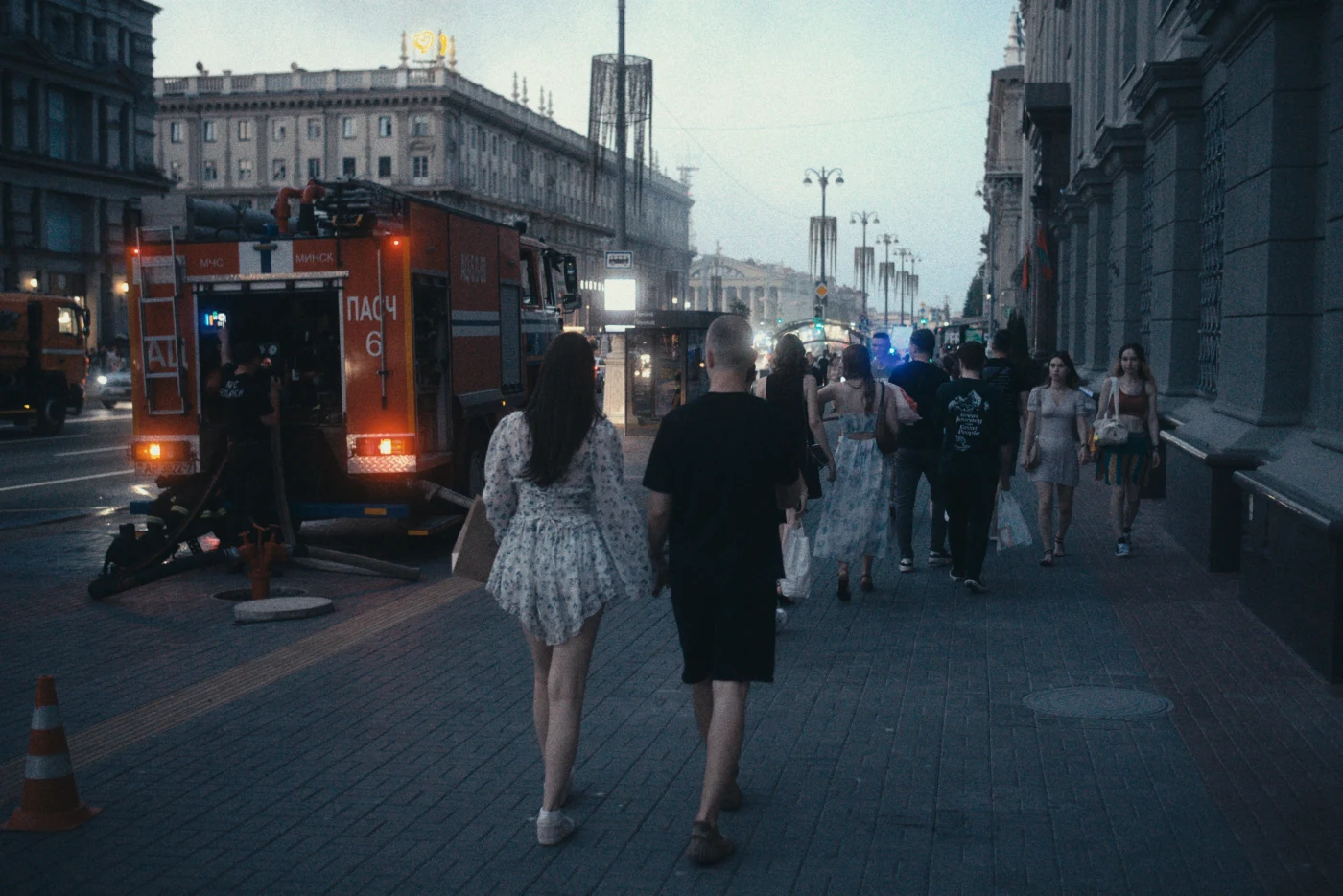 People walking along the street next to a fire truck. Buildings and evening lights are visible in the background.