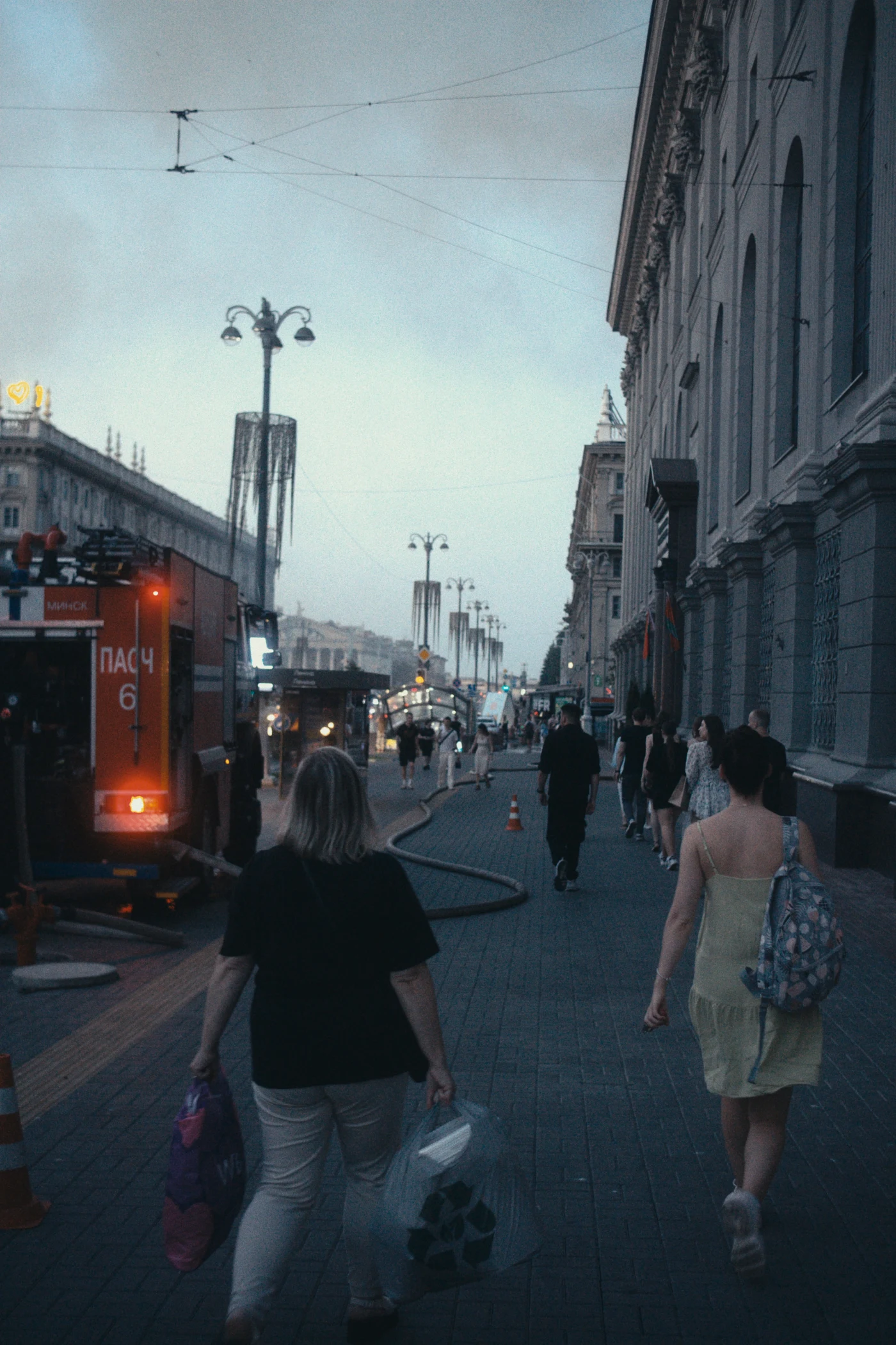 A pedestrian street in the evening, with people walking past a fire truck and shops.