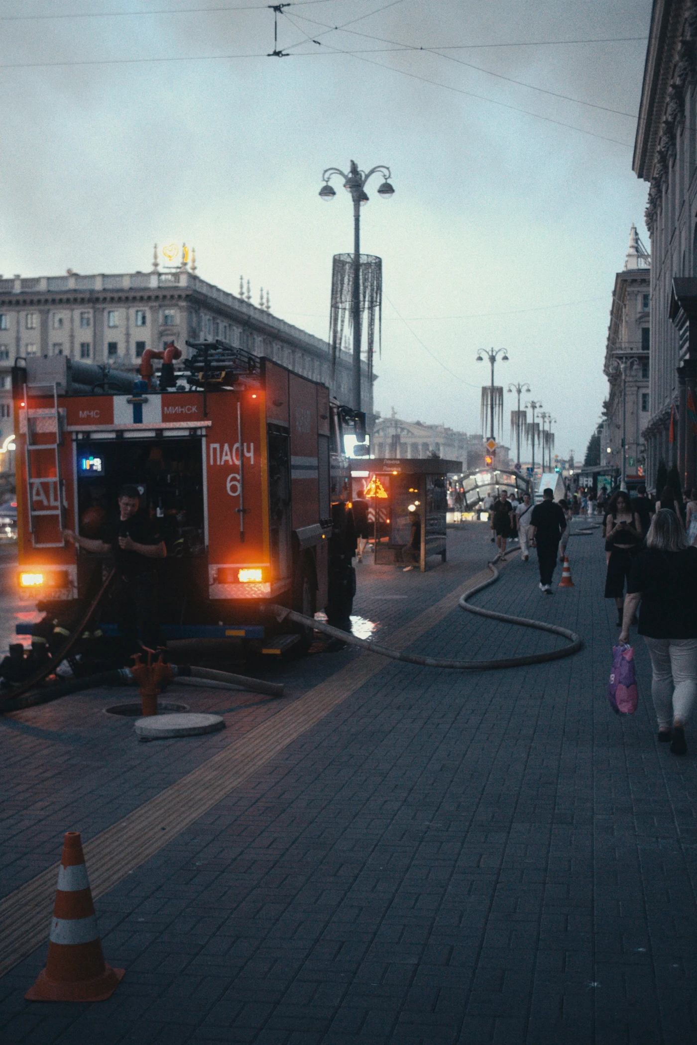 A fire truck on the street, with people walking past, fire extinguishers and hoses visible. Buildings and evening lighting are visible in the background.