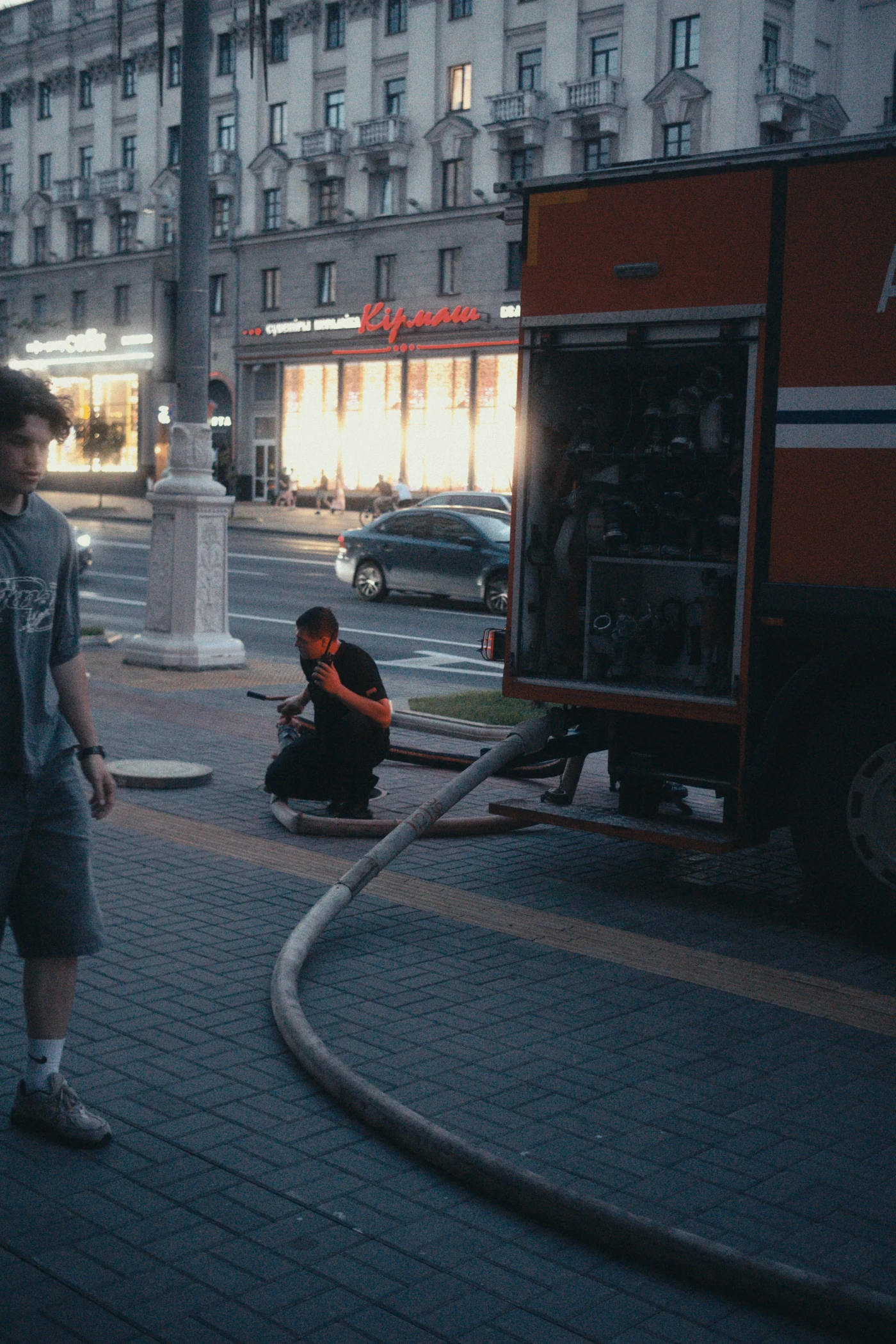A fire truck on the street, with one person standing nearby and another sitting on the ground using a phone. Buildings and shop windows are visible in the distance.