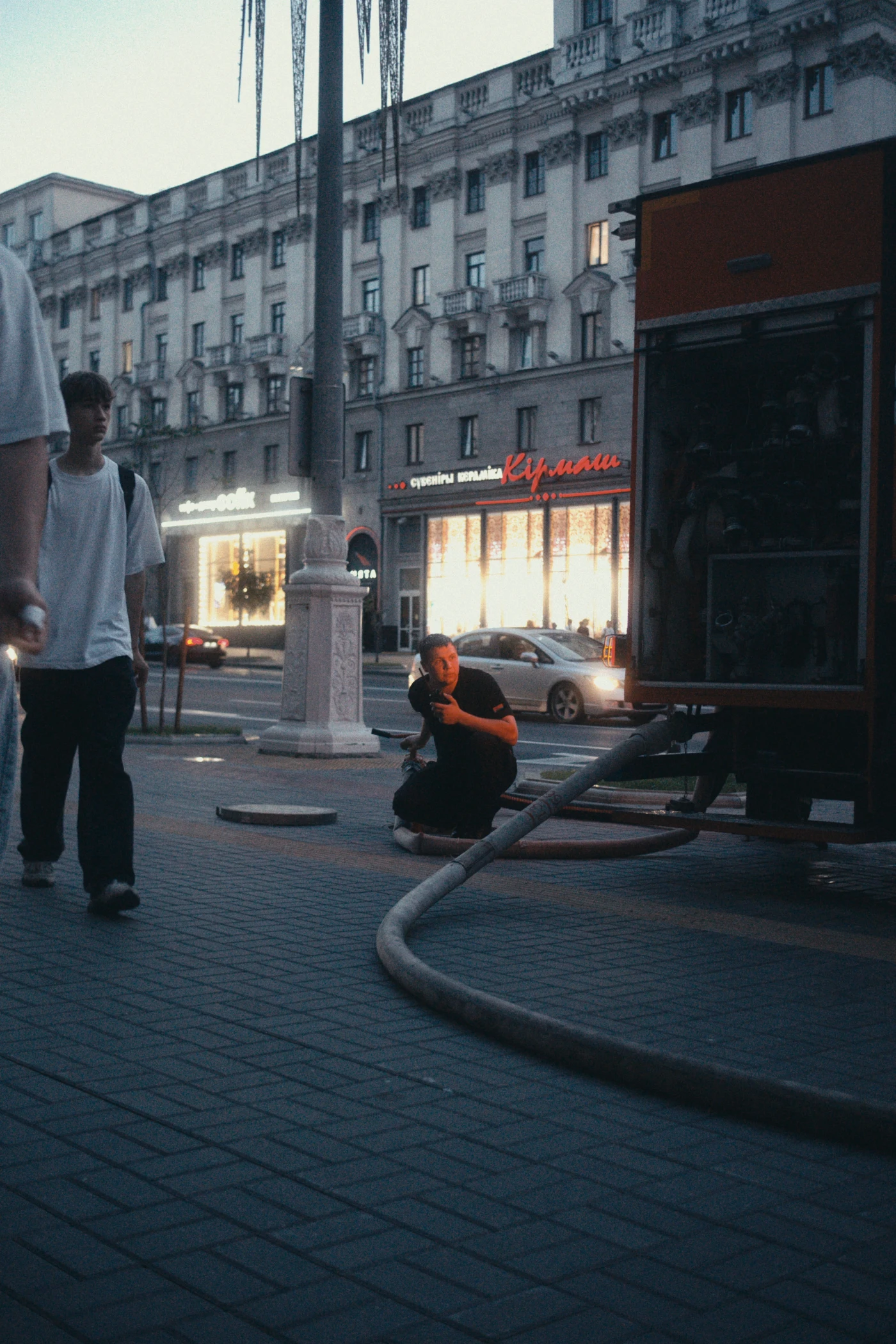 An evening street, with people walking past and a firefighter working with a hose near the truck. Shop windows glow in the distance.