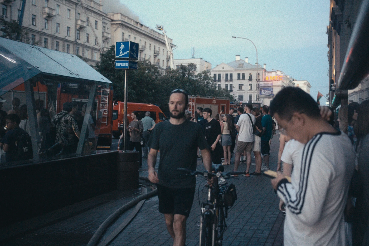 A crowded street, with a man walking past with a bicycle and other people standing and looking at their phones.