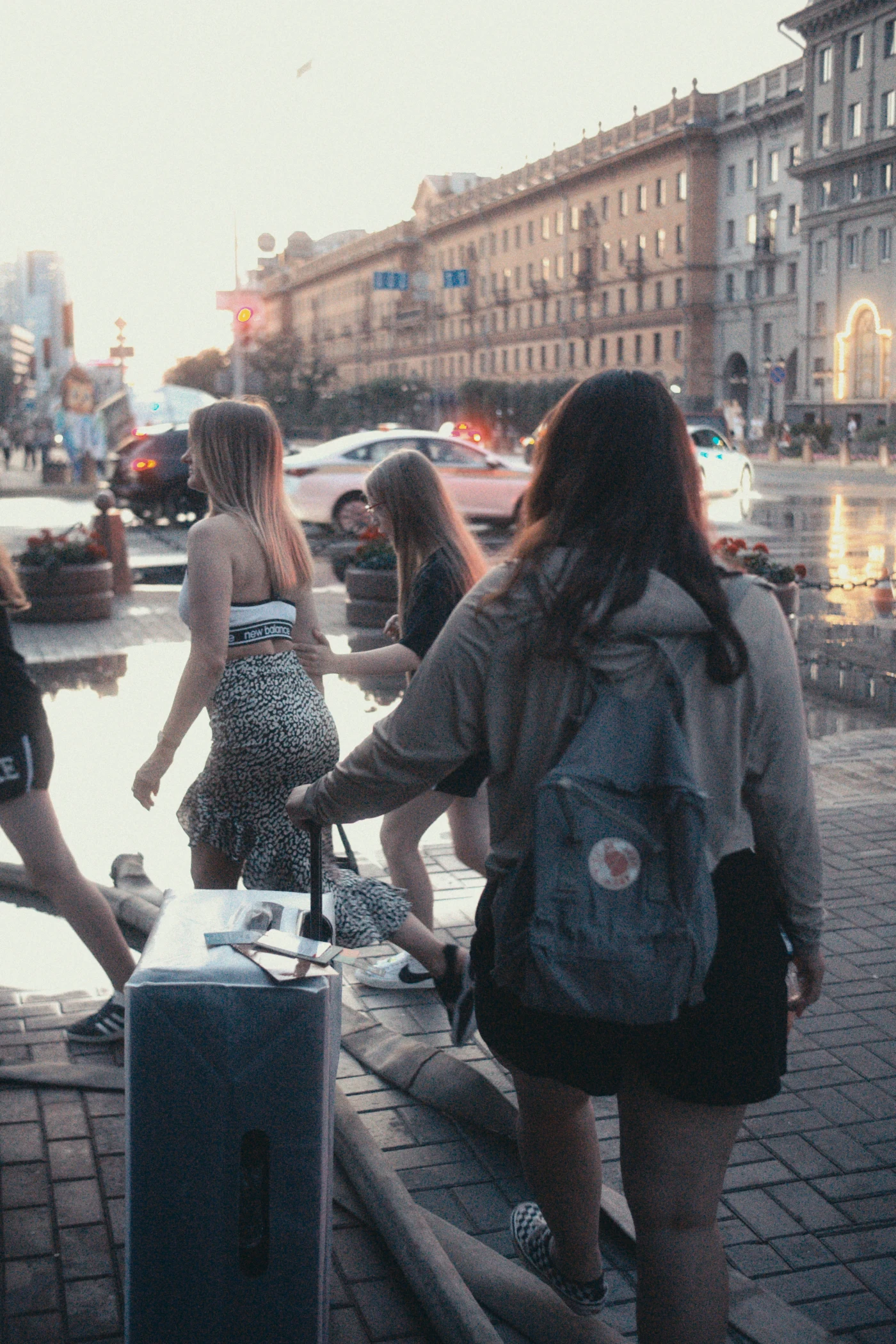 A group of girls on the street, one with a suitcase, in the evening city, with reflections in puddles.