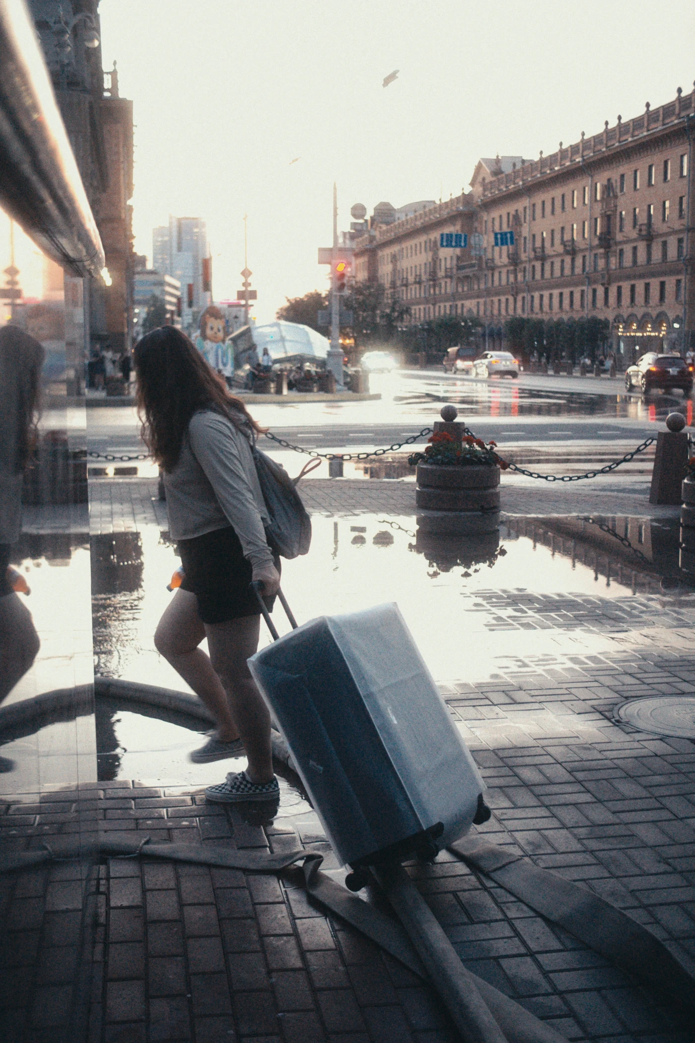 A girl with a suitcase walking down the street, with reflections of buildings and puddles on the asphalt. City views are visible in the distance.