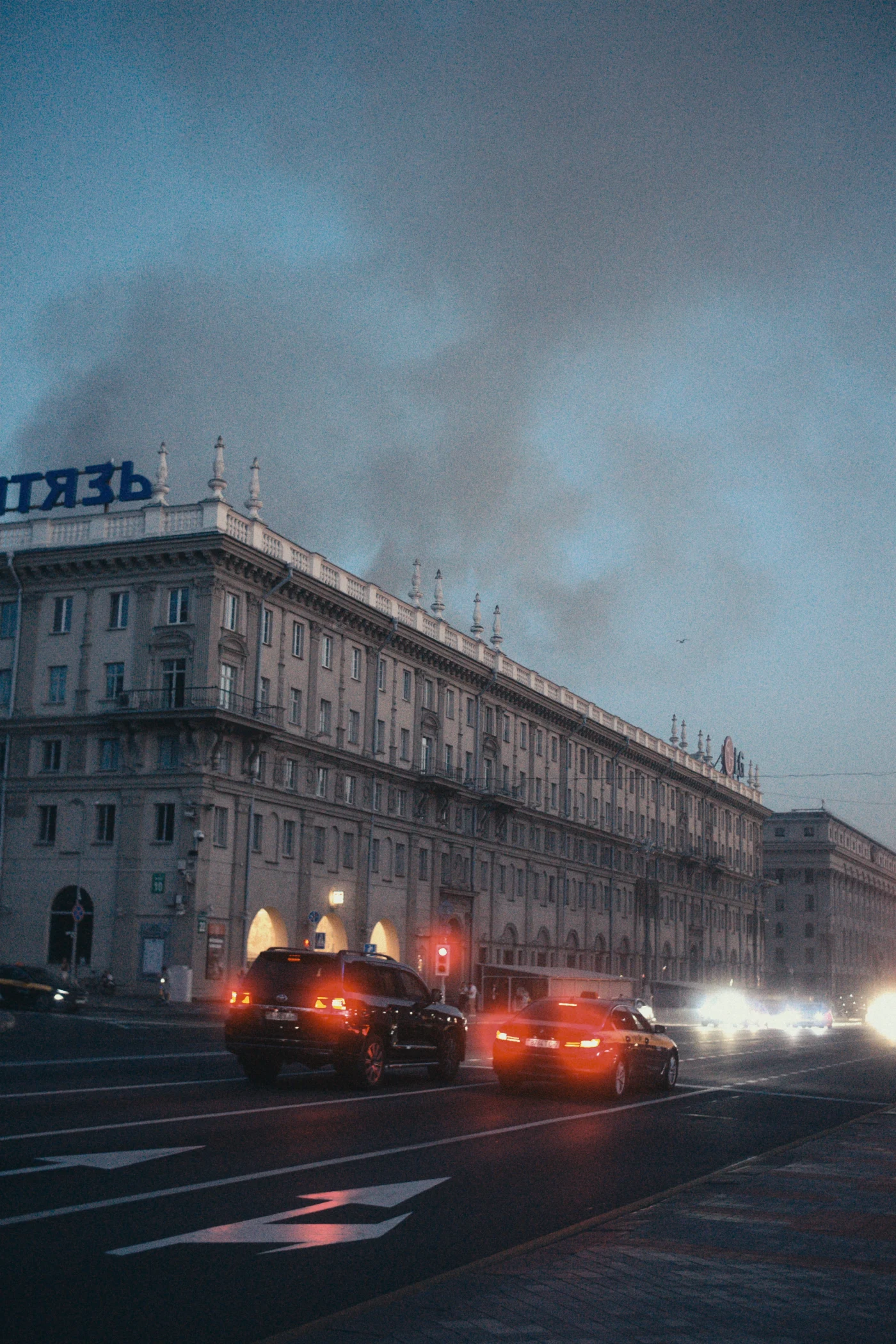 A street with car traffic, evening lighting, and a cloudy sky. The word “ГИТАЖЬ” is visible on a building.