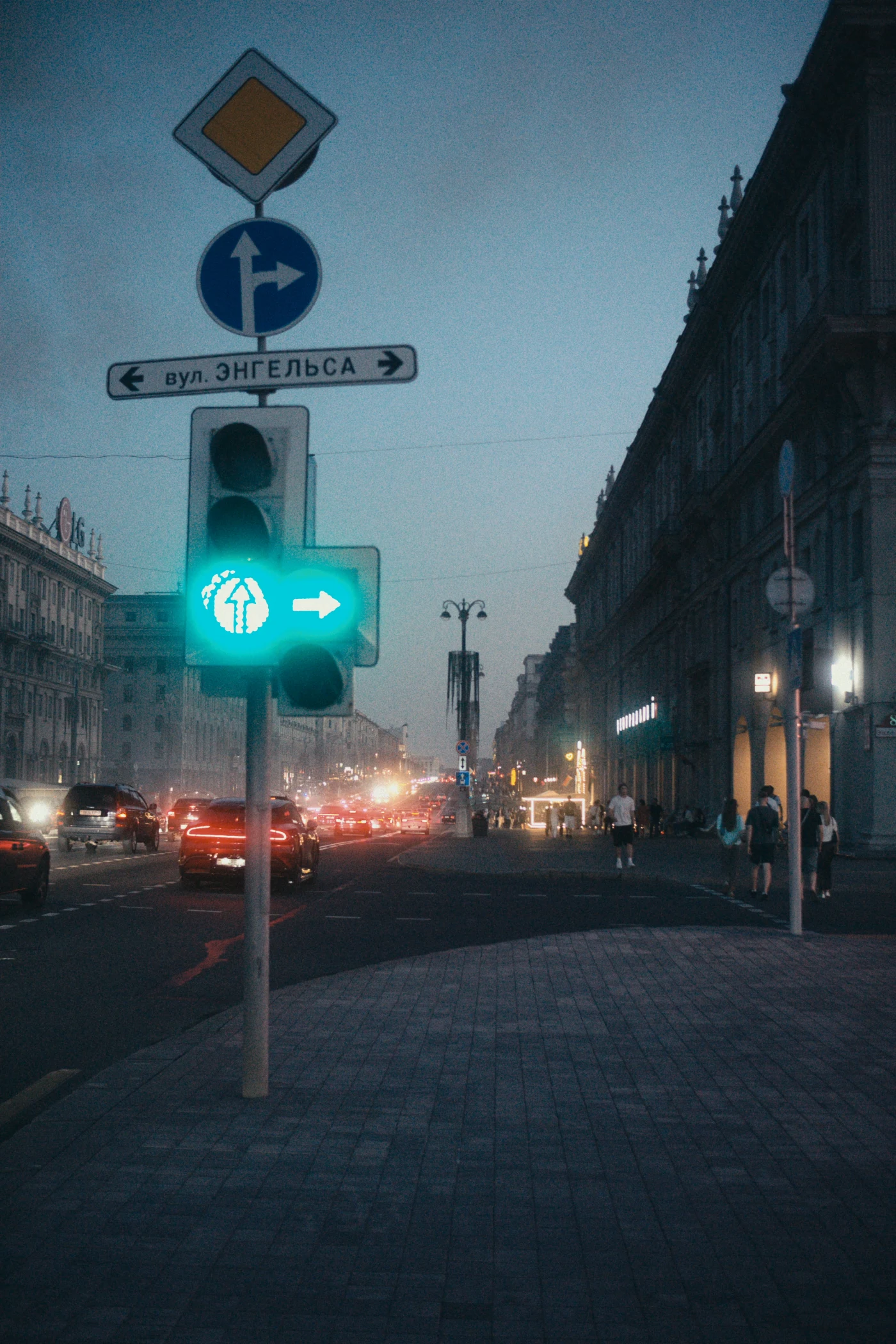 A street with a green traffic light and road signs, evening lighting, cars, and pedestrians on the sidewalk.