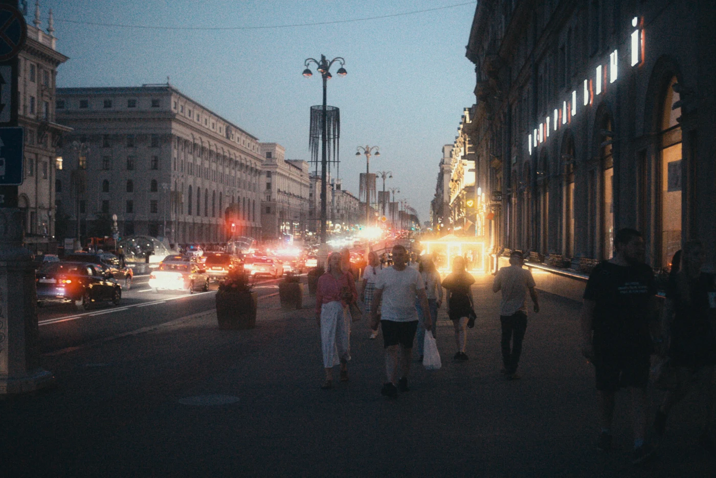 A nighttime cityscape with people on the sidewalk and car traffic, with illuminated buildings along the street.