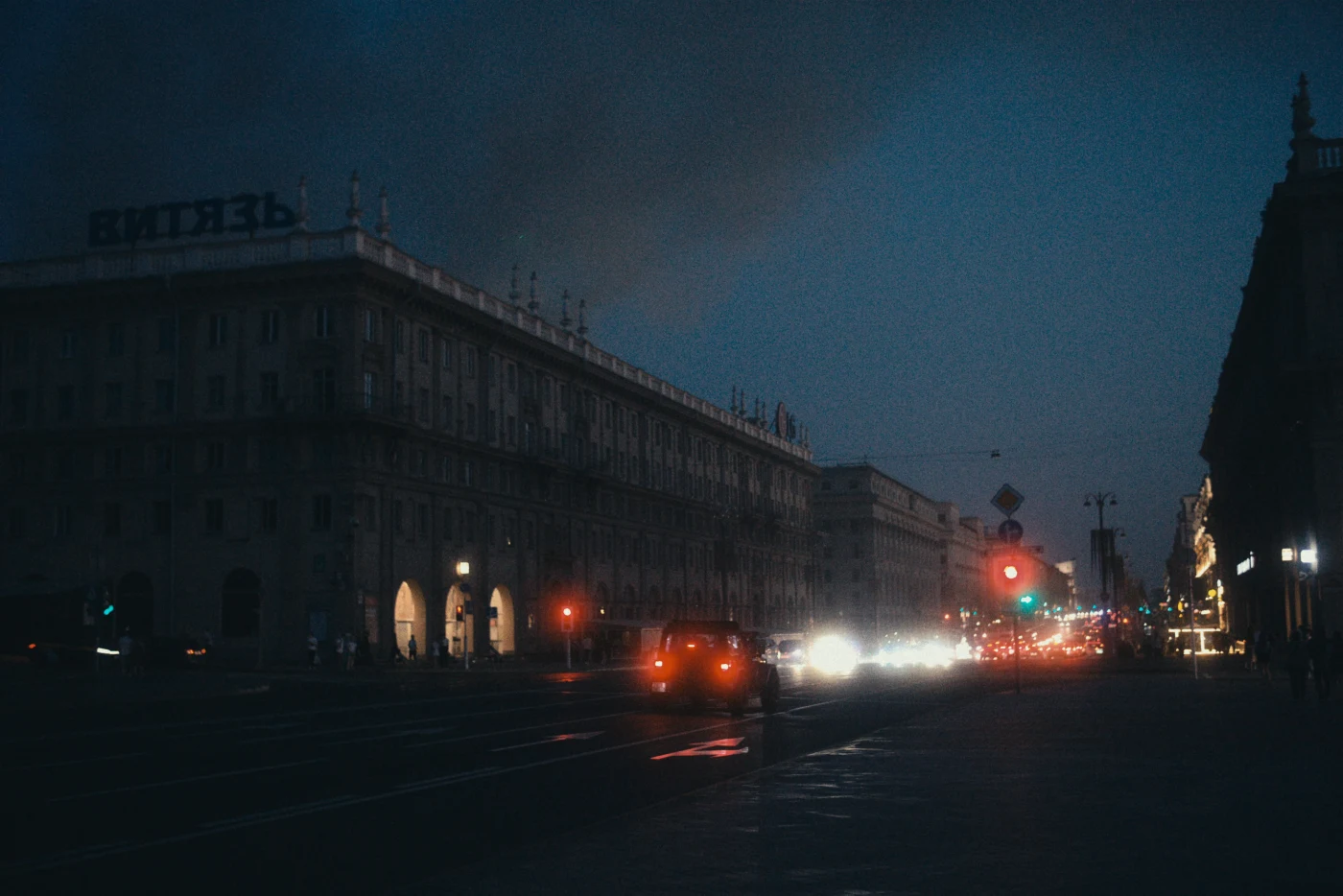 A nighttime cityscape with illuminated buildings and moving cars. The word “ВИТЕЗЬ” is visible on the roof.