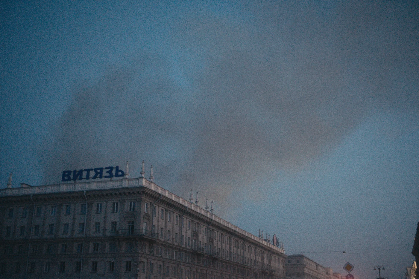 The word “ВИТЯЗЬ” is written on a building, with smoke rising above it against the evening sky.