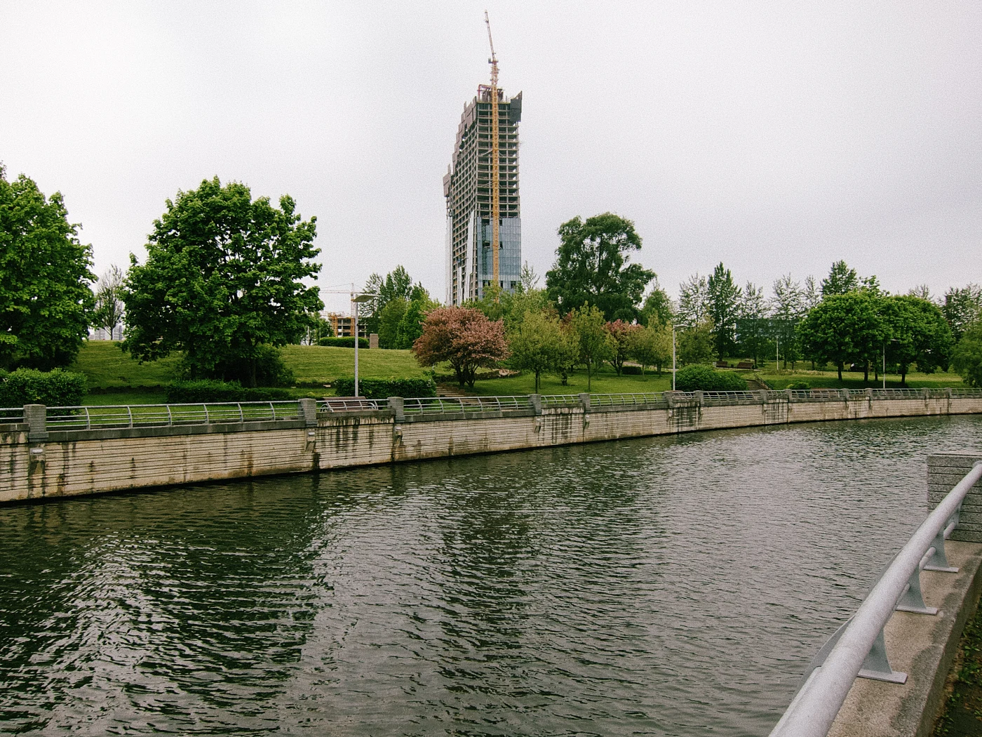 A river with green trees along the bank and a building under construction against a gray sky.
