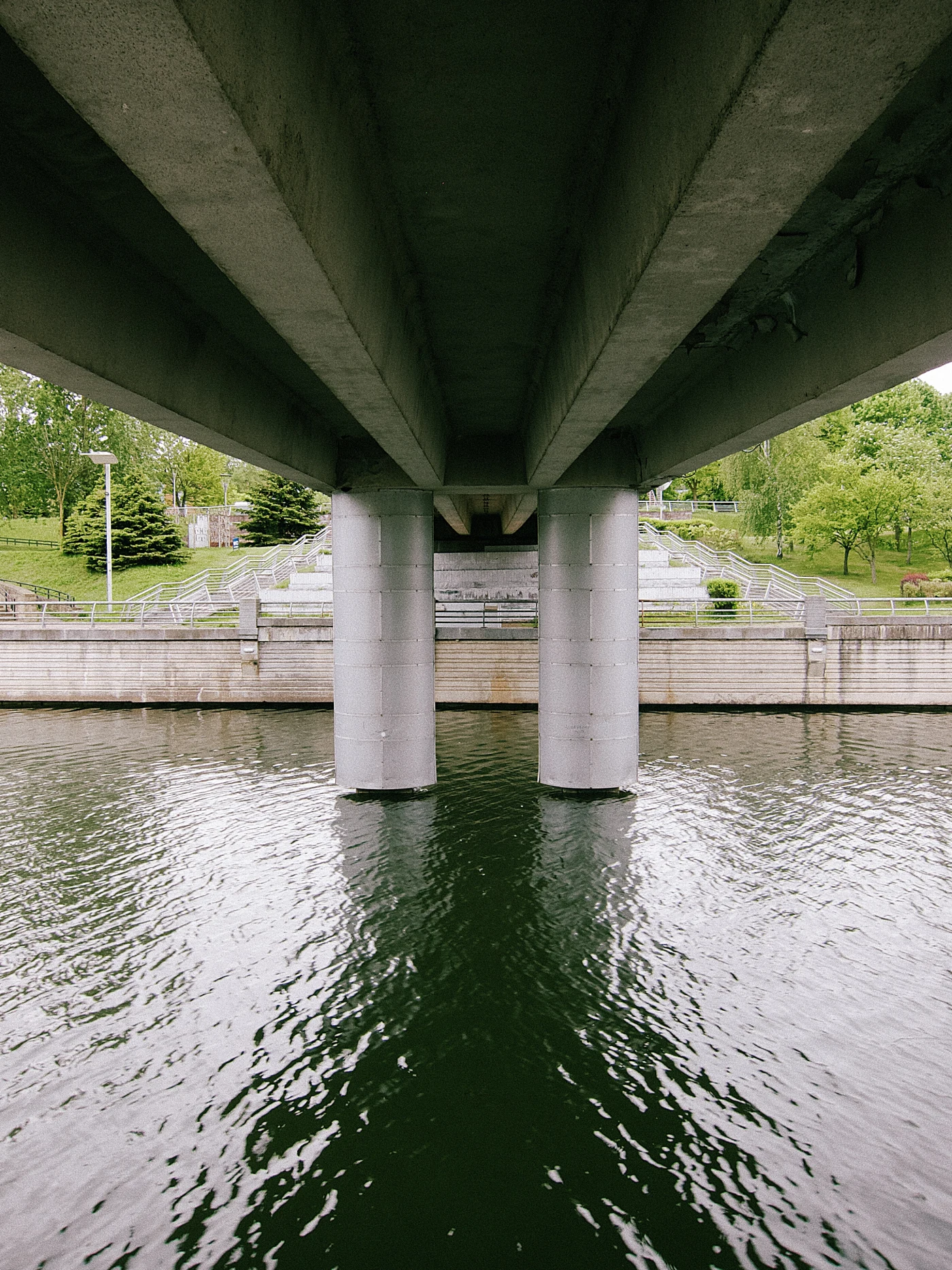 Bridge supports above the water, green trees and stairs on the bank. A calm water surface.