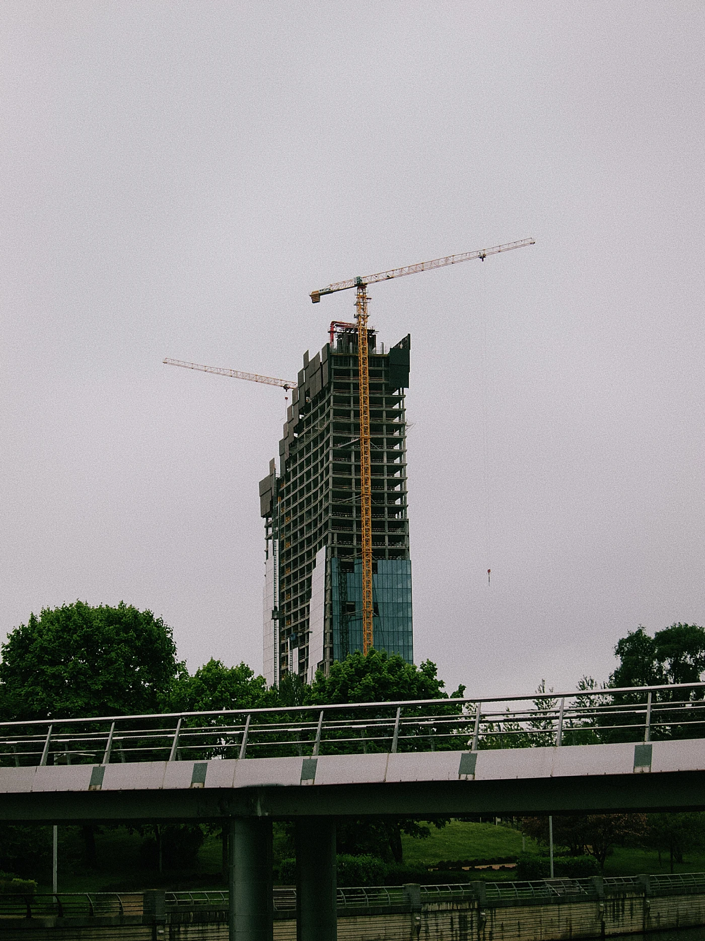 A skyscraper under construction with cranes against a cloudy sky and green trees.