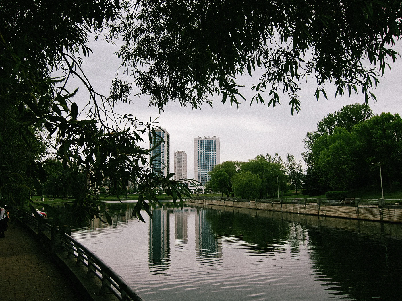An urban landscape: a river with reflections of buildings and green trees on the bank. The sky is cloudy.