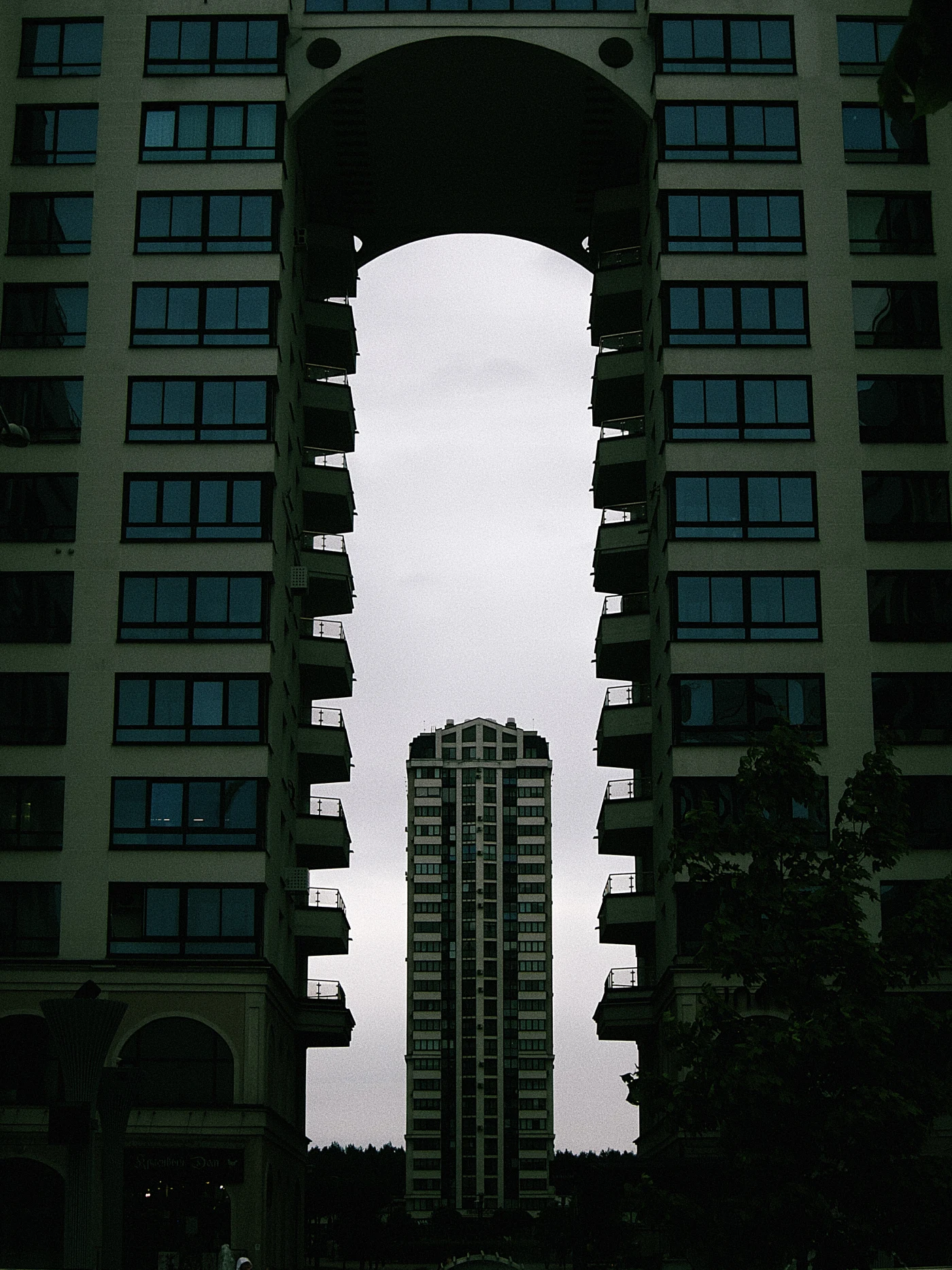 An architectural composition: two buildings with an arch, with another skyscraper visible in the center against a gray sky.