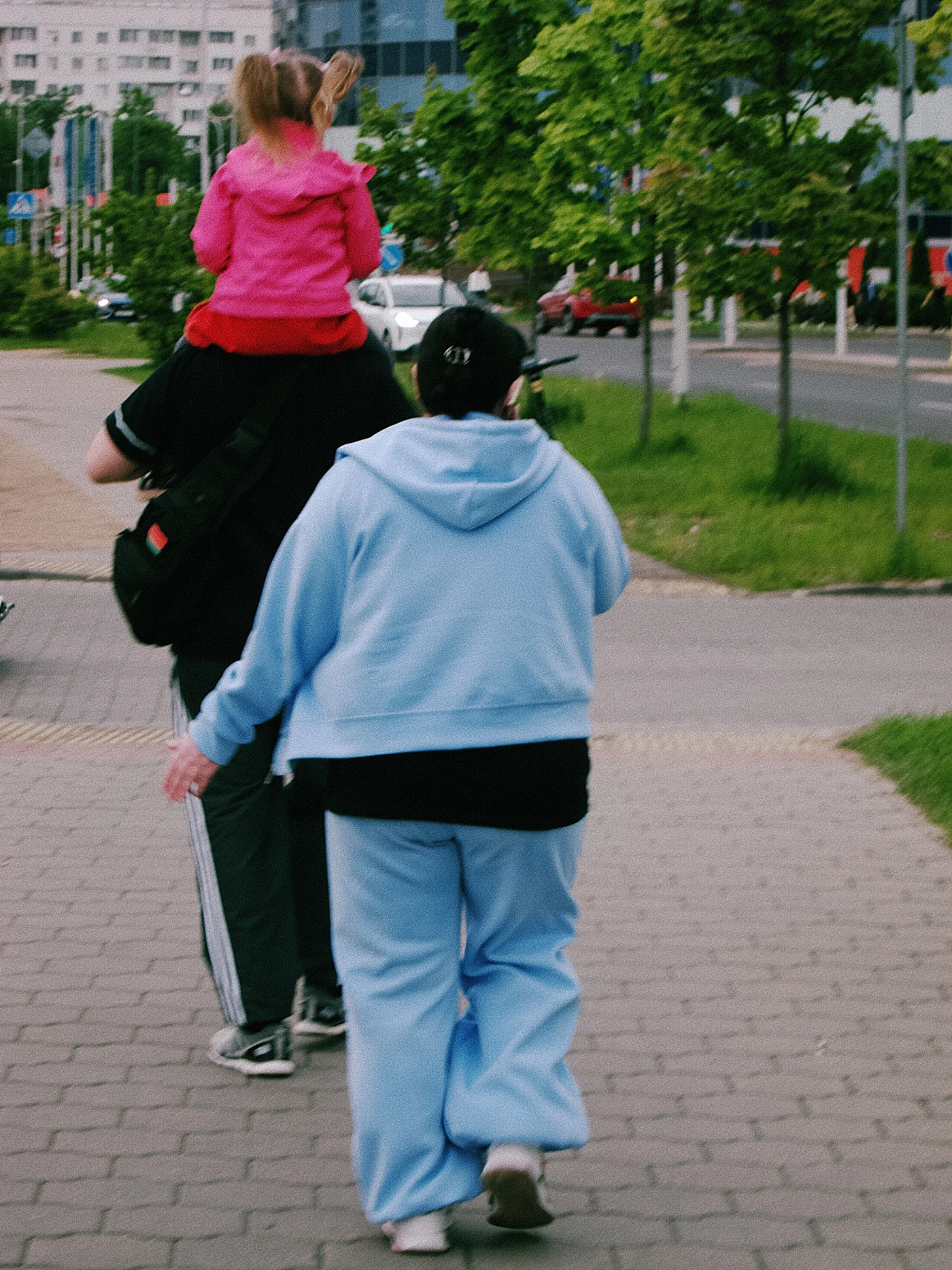 A woman in a blue suit and a man carrying a girl on his shoulders walk along the sidewalk. Greenery and city buildings around them.