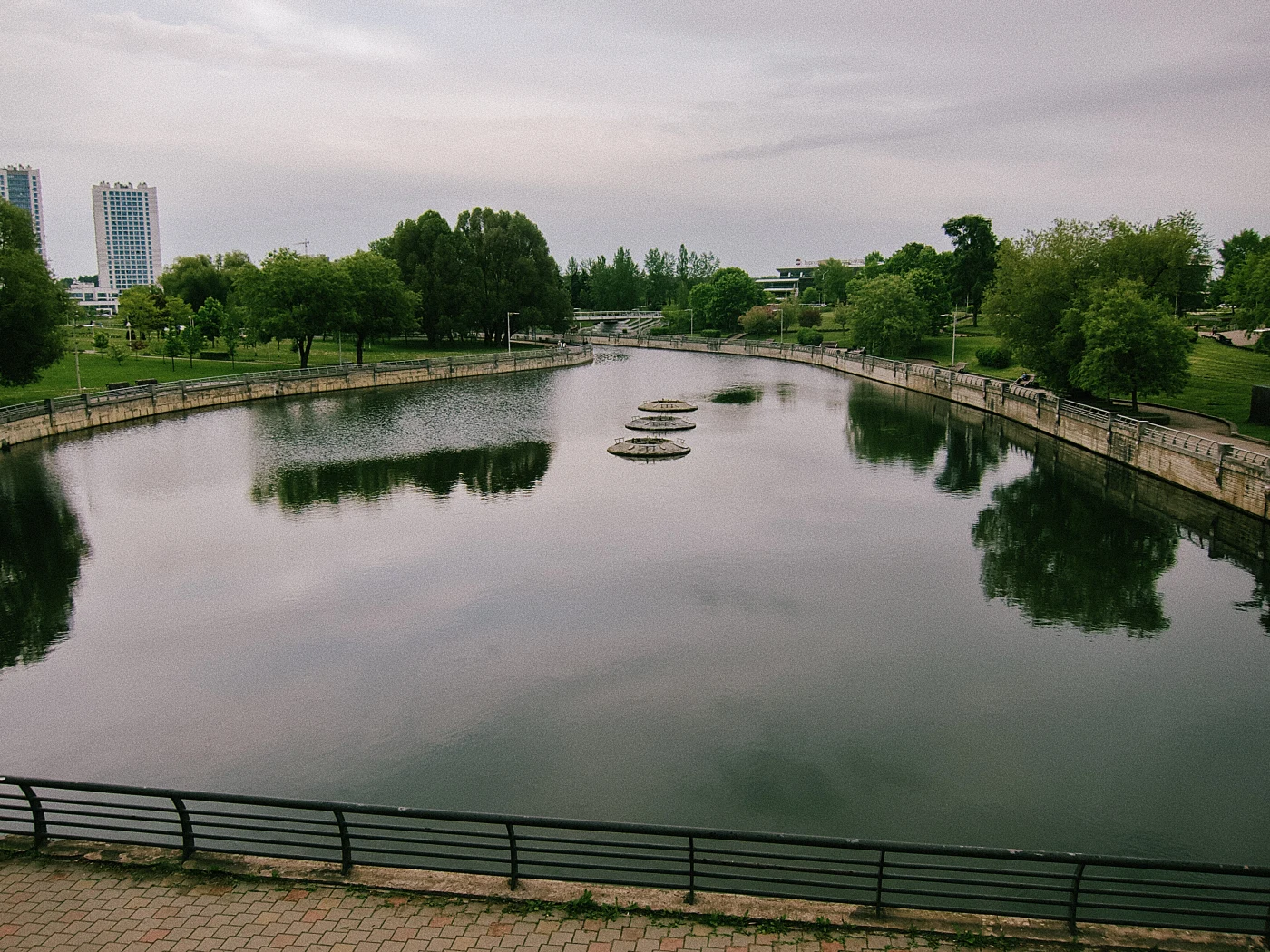 A calm river with reflections of trees and buildings, with a walking area and green plantings along the bank.