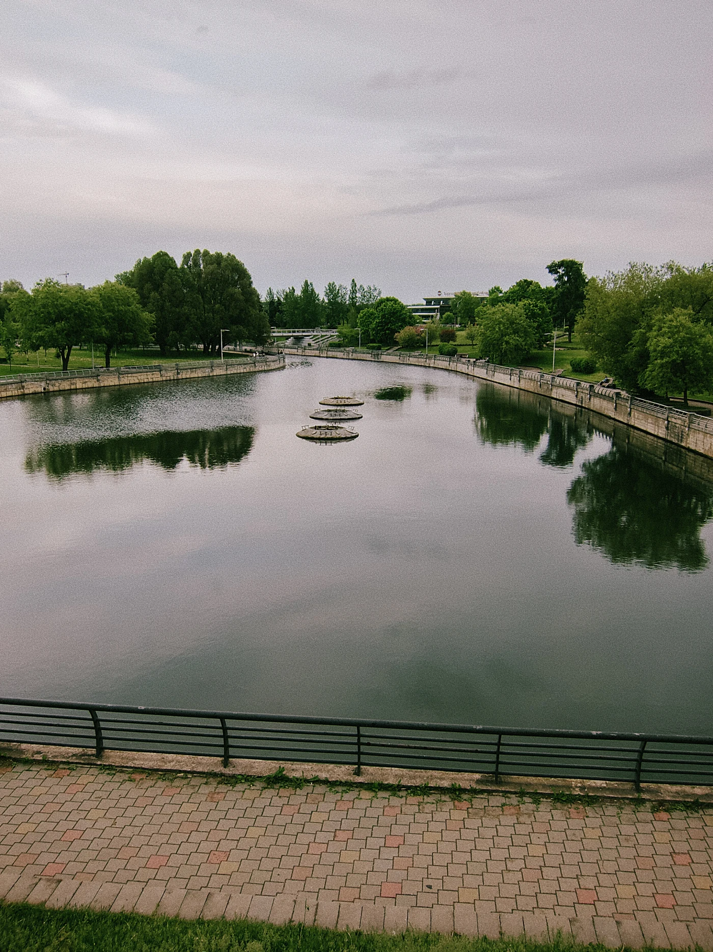 A calm river with reflections of trees and stone islands on the water surface. A gray sky above the horizon.