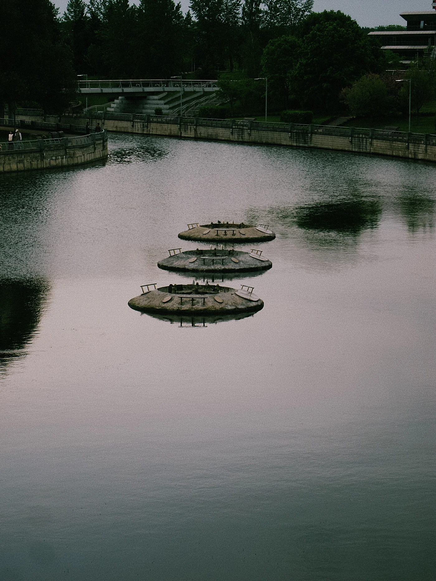 Three round platforms are visible on the calm river water, surrounded by green trees and the bank.