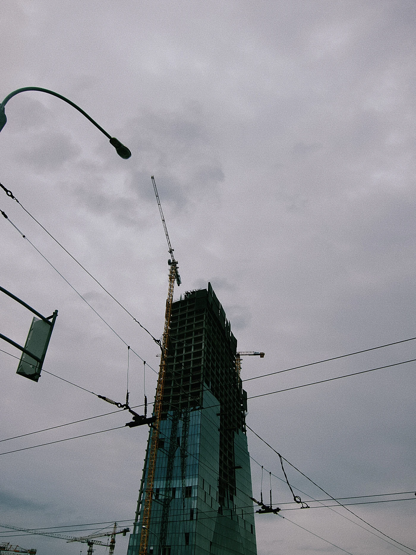 A skyscraper under construction with cranes against a cloudy sky and power lines.