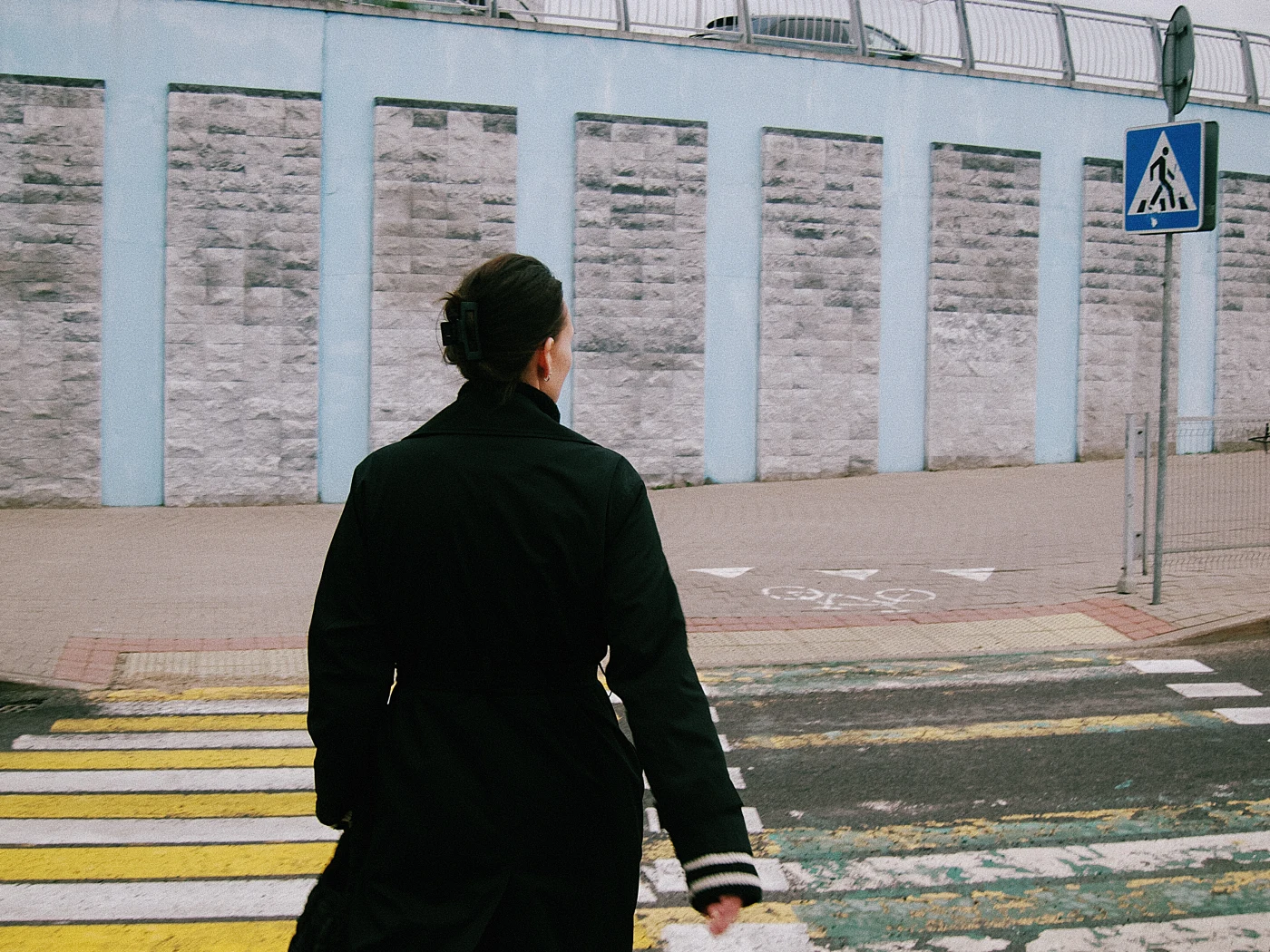 A woman in a black coat walks across a pedestrian crossing, with a gray wall and road sign in the background.