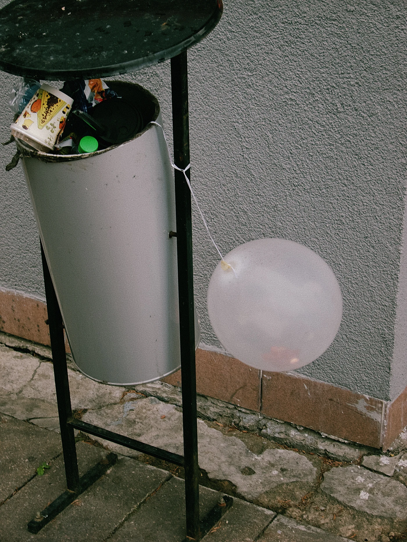 A trash bin with waste and a transparent balloon tied to it. A gray wall.