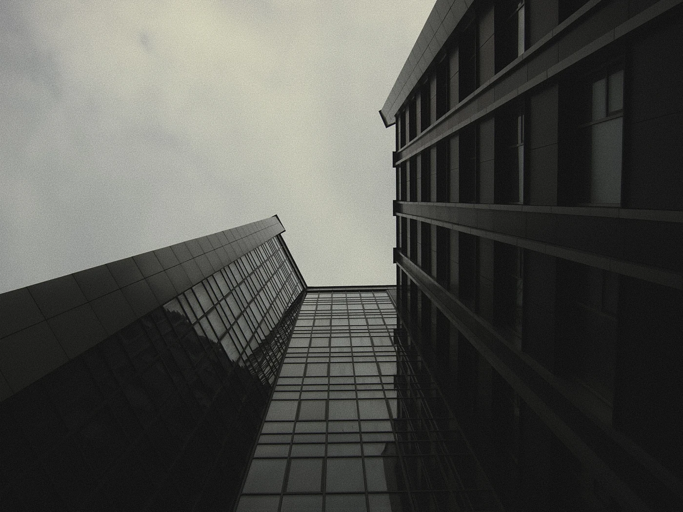 A low-angle view of tall buildings, gray sky and reflections on glass facades.
