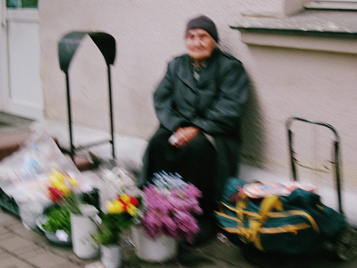 An elderly woman in dark clothing sits on the sidewalk next to flowers and bags.
