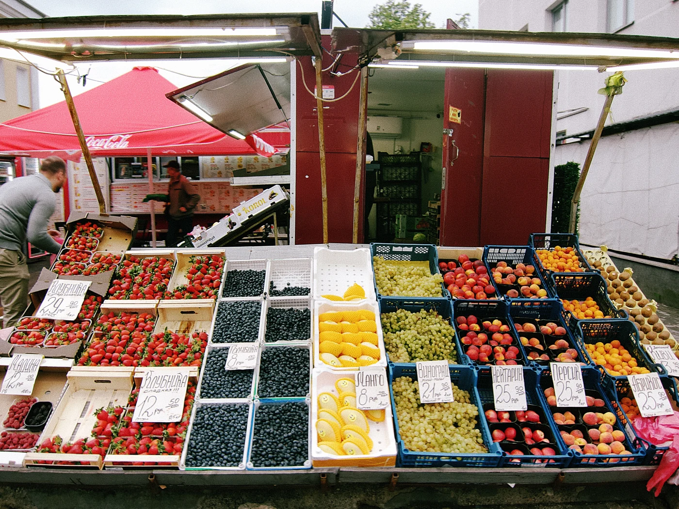 A bright fruit market: strawberries, grapes, peaches, apricots and citrus fruits in baskets, with prices on signs.