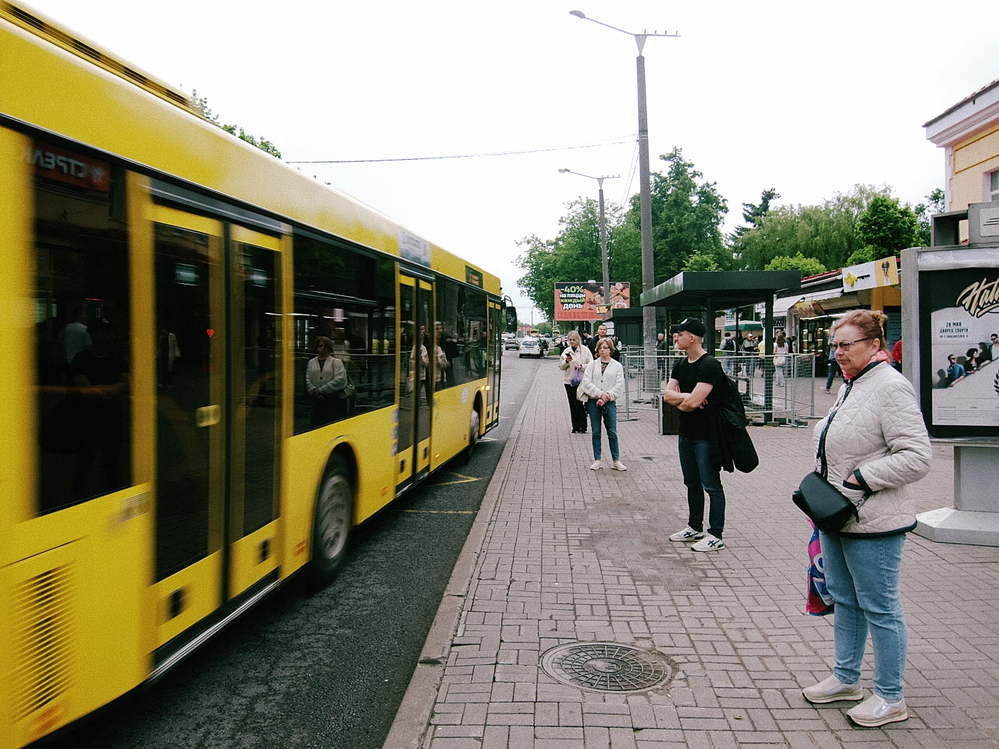 A yellow bus passes a stop where people are waiting for transport. The weather is cloudy.