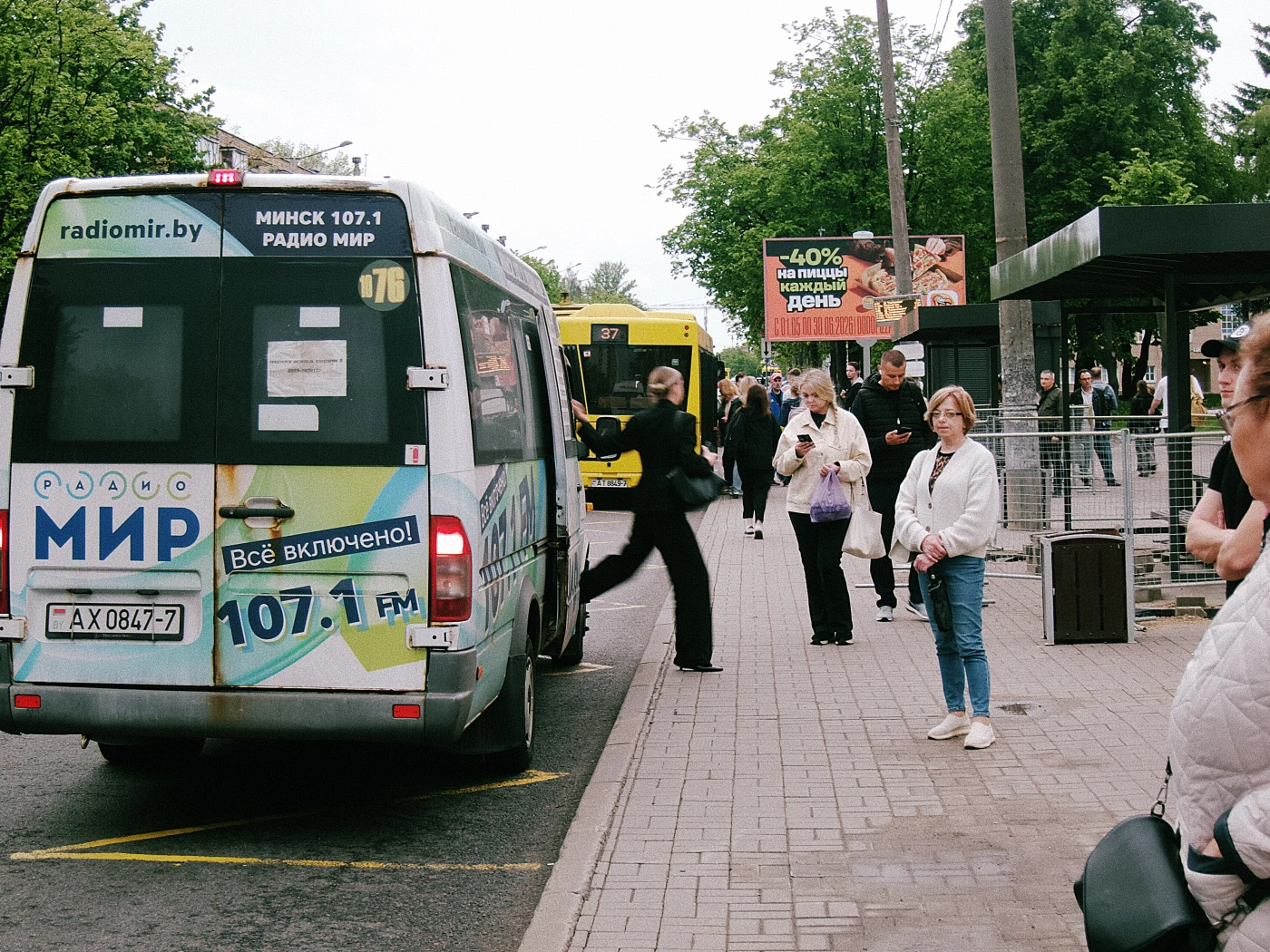 A bus with an ad for MIR radio at a stop, people waiting for transport, one passenger boarding the bus.