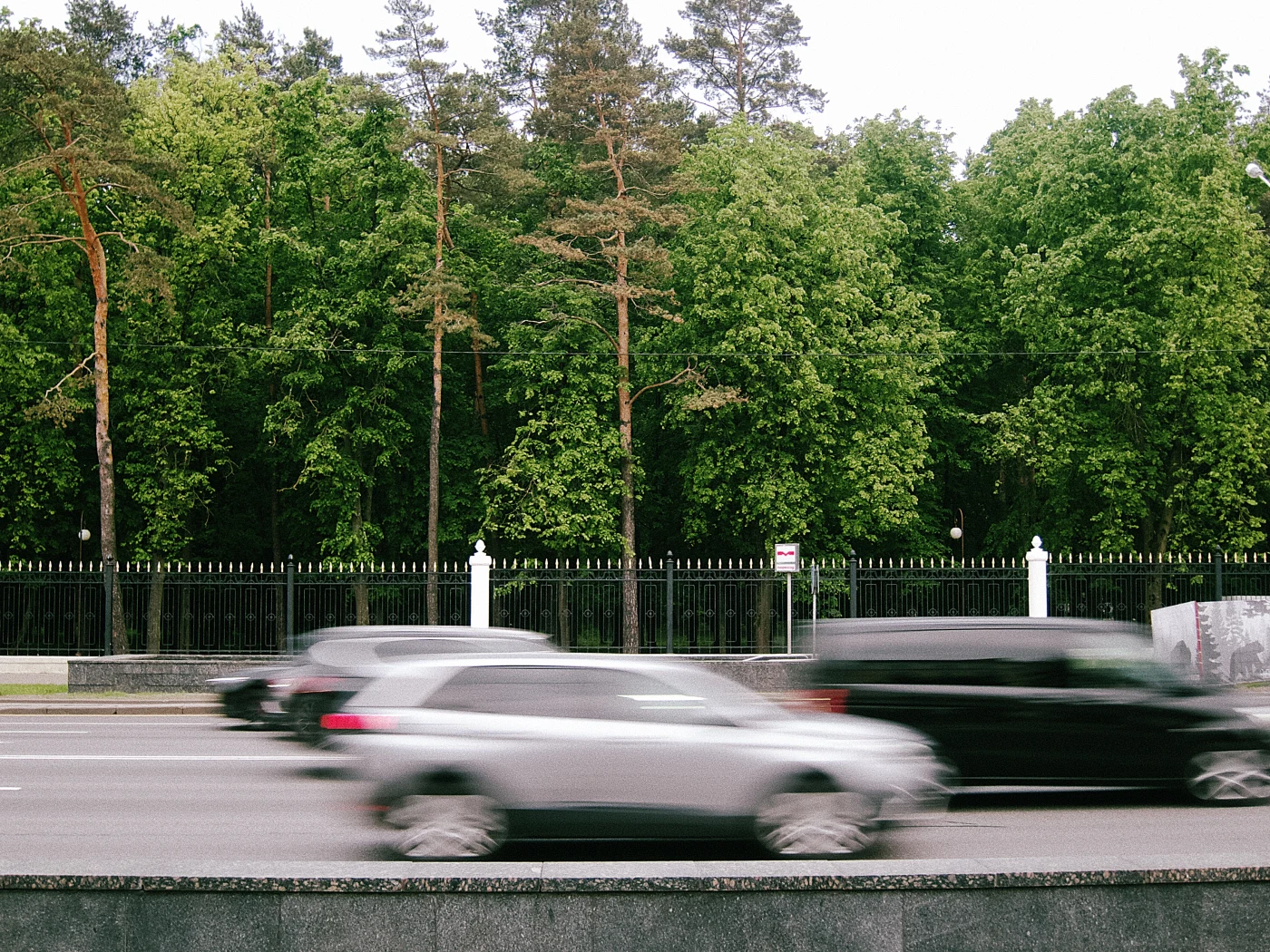 Blurred cars against a background of green forest and a fence.