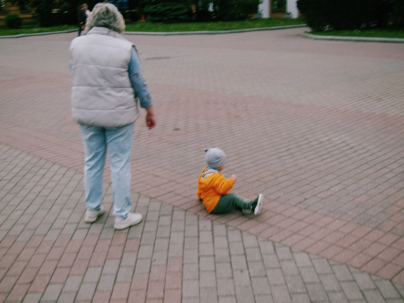 A woman in a vest walks across the tiles, while a toddler in an orange sweater sits on the ground.