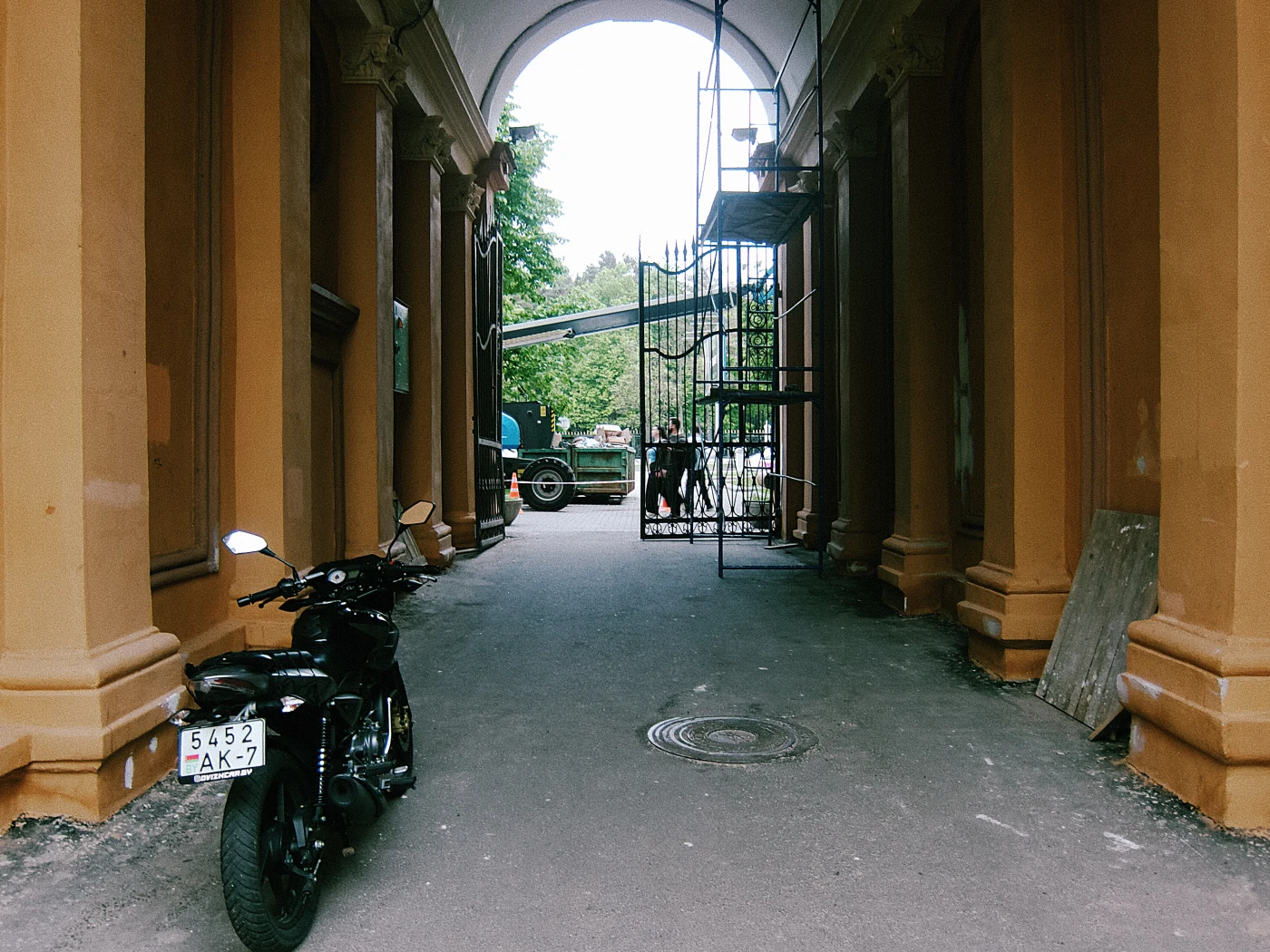 A motorcycle stands under an arch, with workers and scaffolding in the distance. A truck is visible on the left.