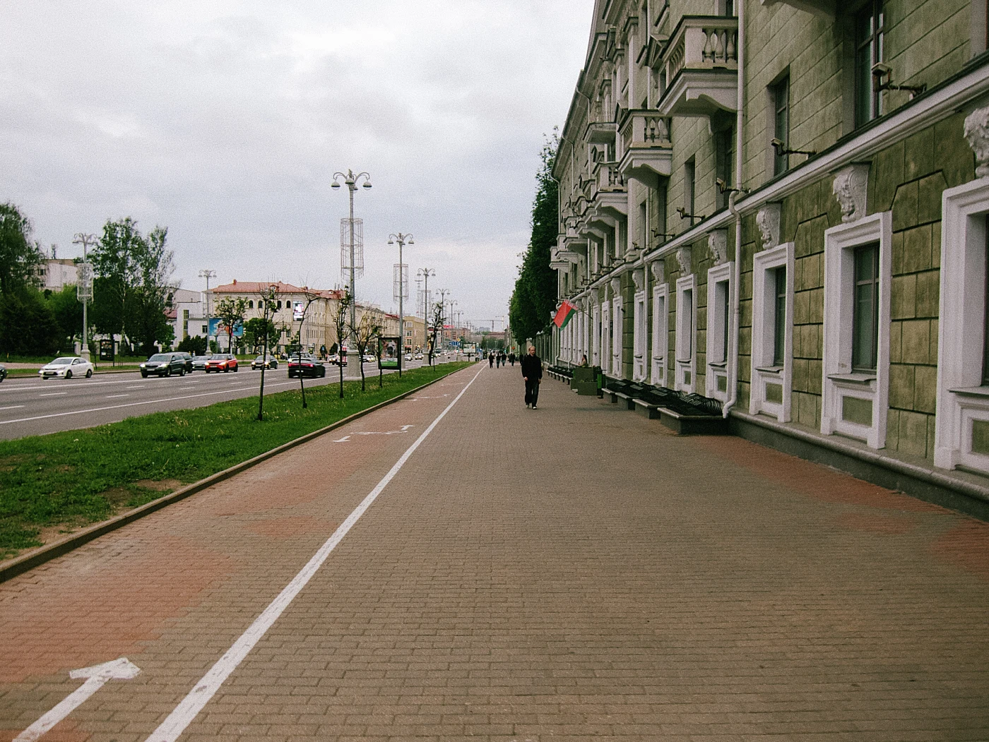 A pedestrian street with a sidewalk lined with trees and buildings, with cars and people visible in the distance.