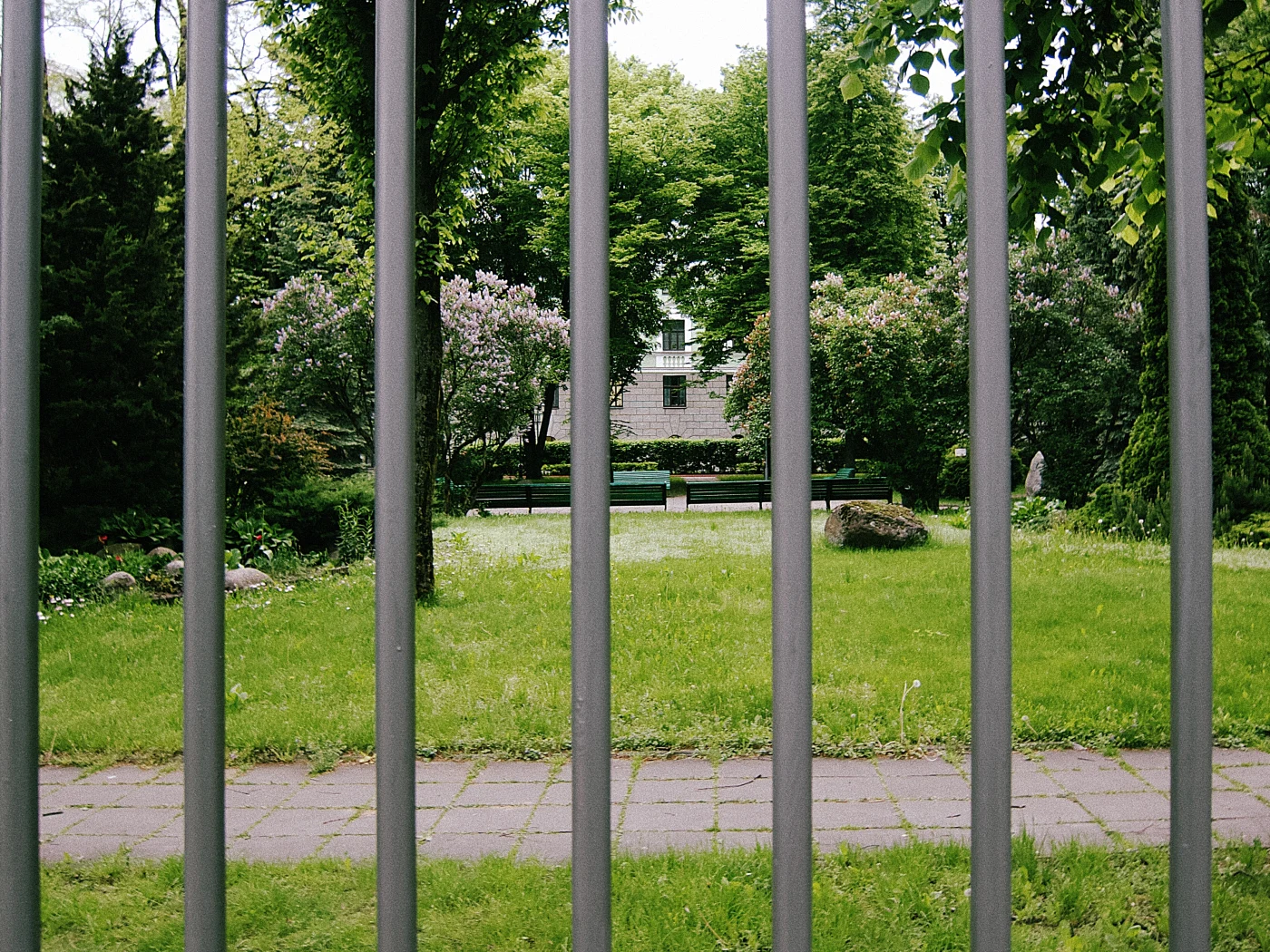 A green park with benches, seen through the metal bars of a fence. A house is visible in the distance.
