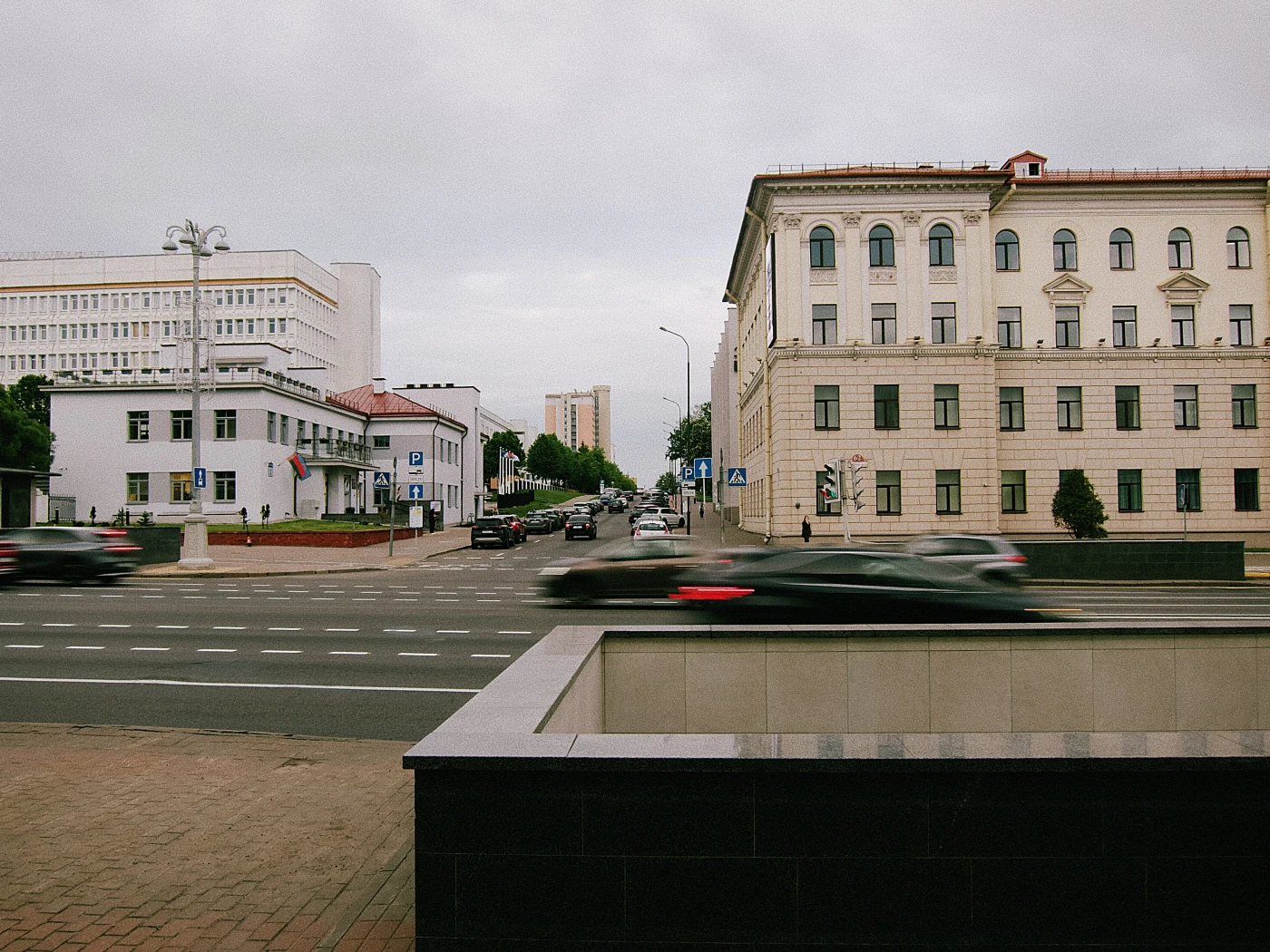 An urban street with cars, buildings and a gray sky. A curb is visible in the foreground.