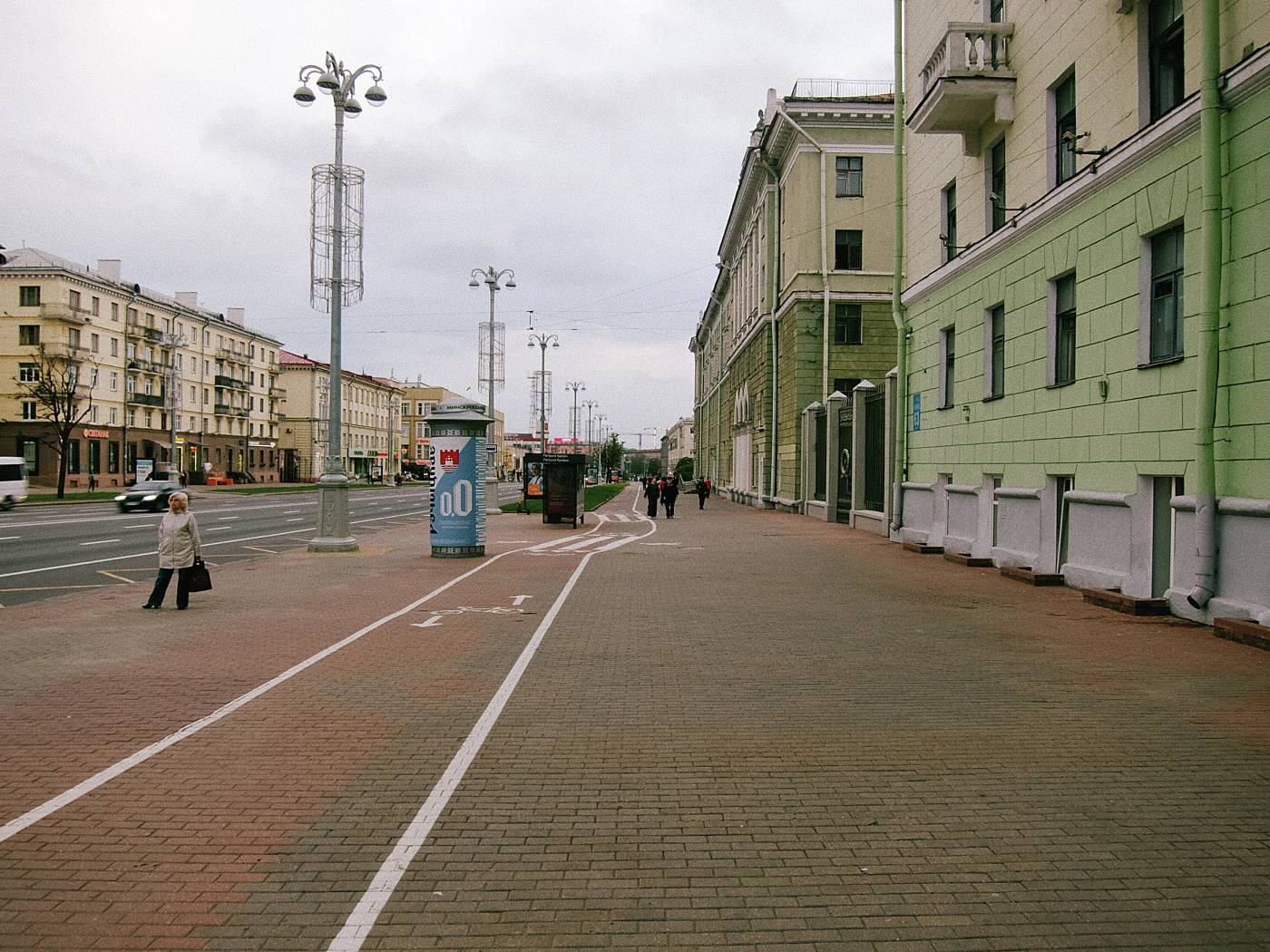 A pedestrian street with green buildings, passersby, a gray sky, with streetlights and billboards visible in the distance.