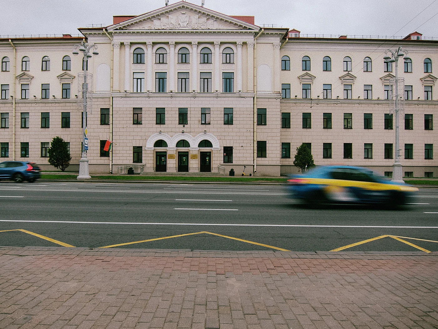 A building with columns and arches, cars passing in the foreground, gray sky.