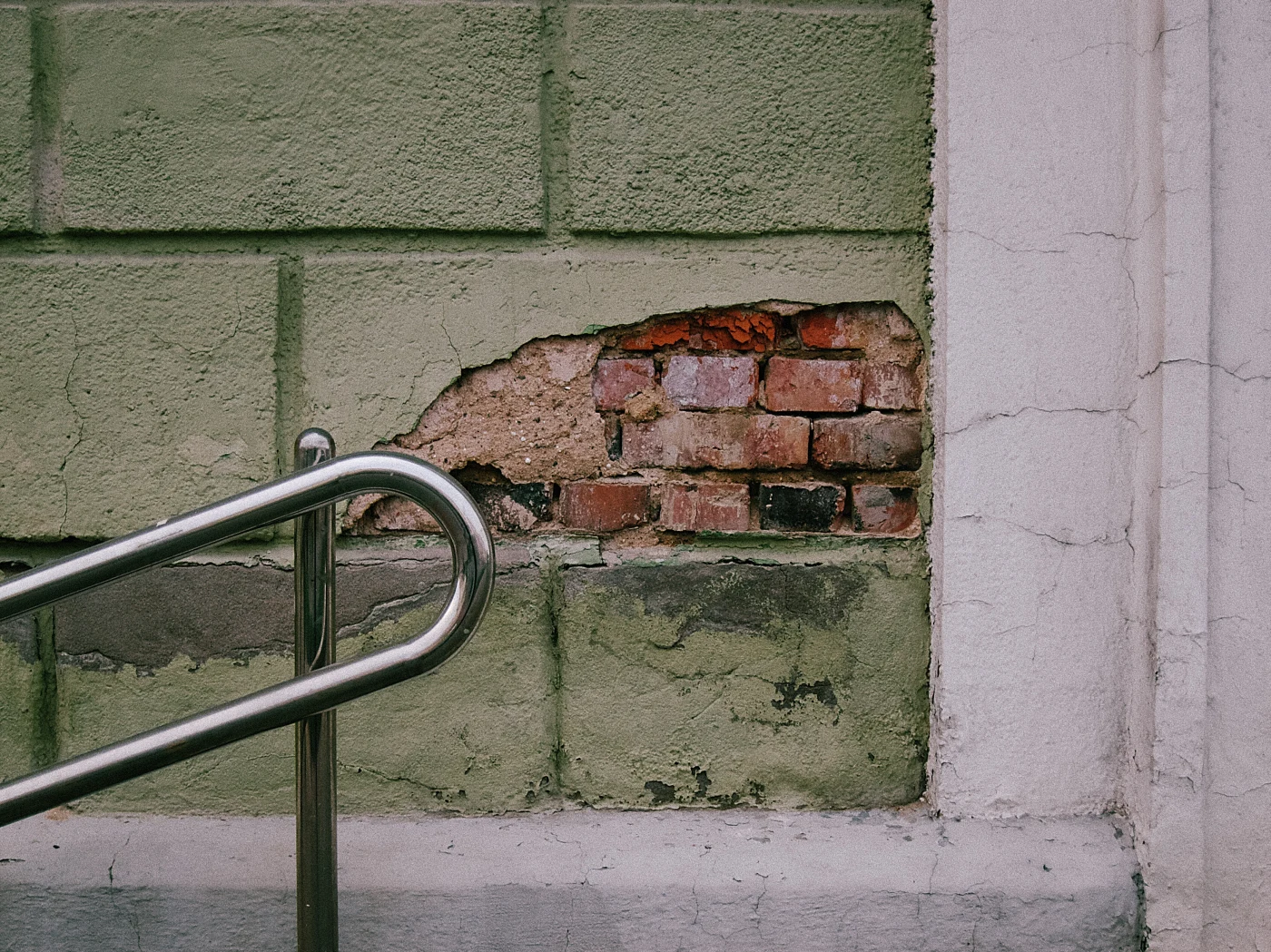 Exposed brick in a wall, next to a metal handrail. A wall with cracks and green paint.
