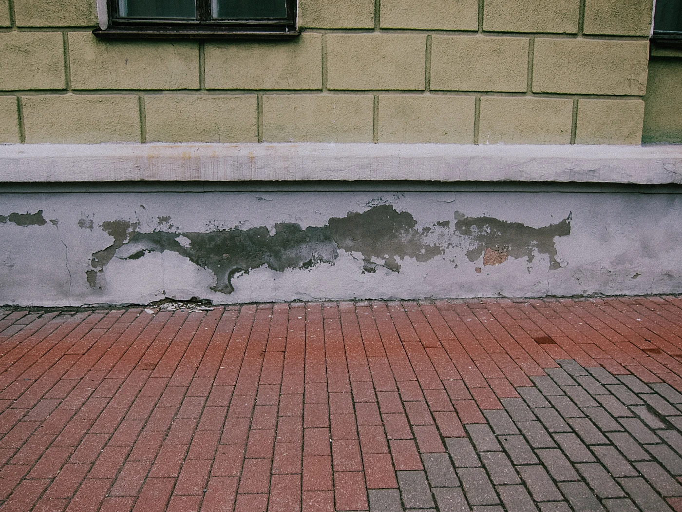 An old wall with peeling paint and brick paving on the sidewalk.