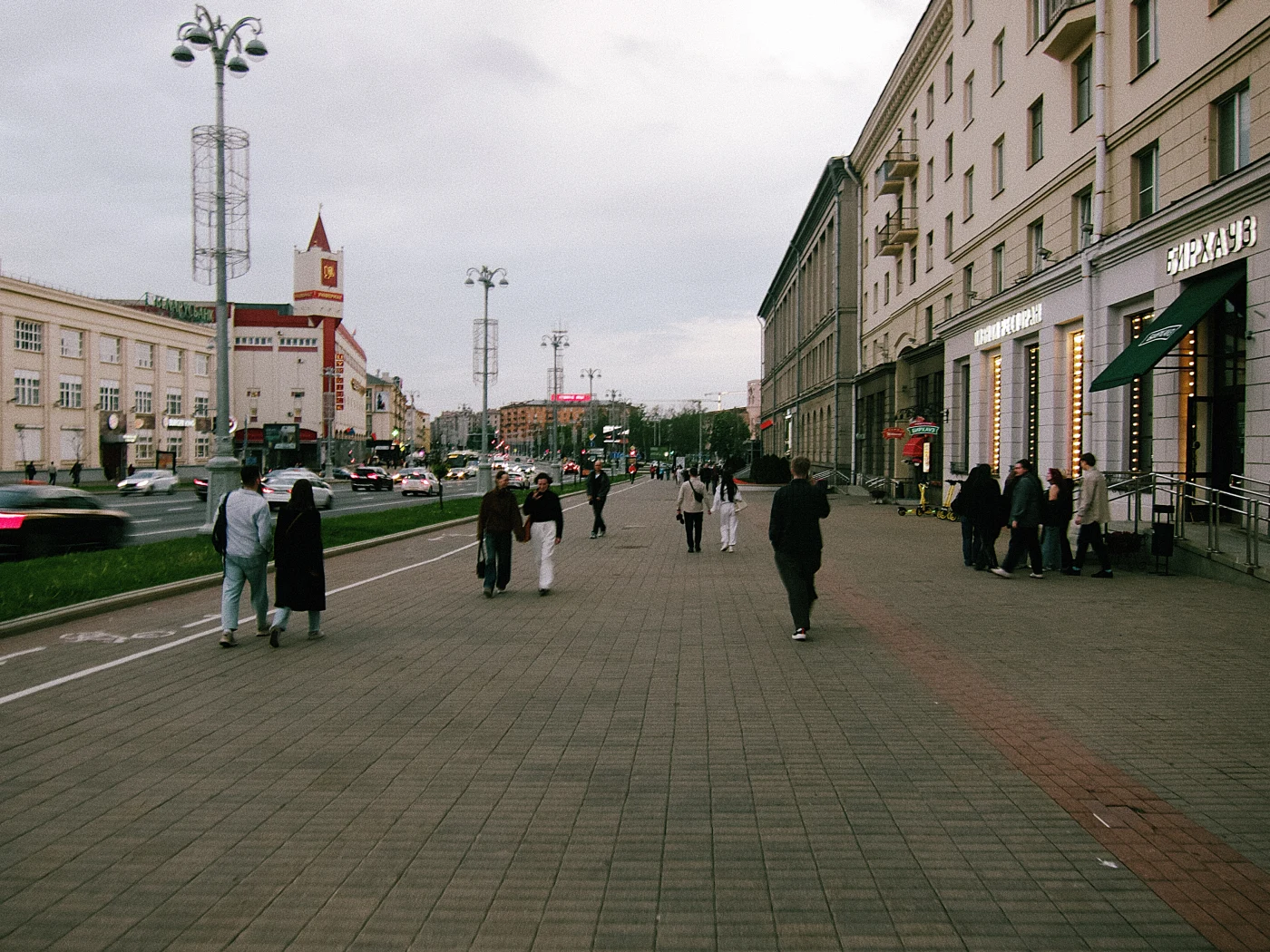 A pedestrian street with people, buildings and evening lighting. A clock tower is visible on the horizon.