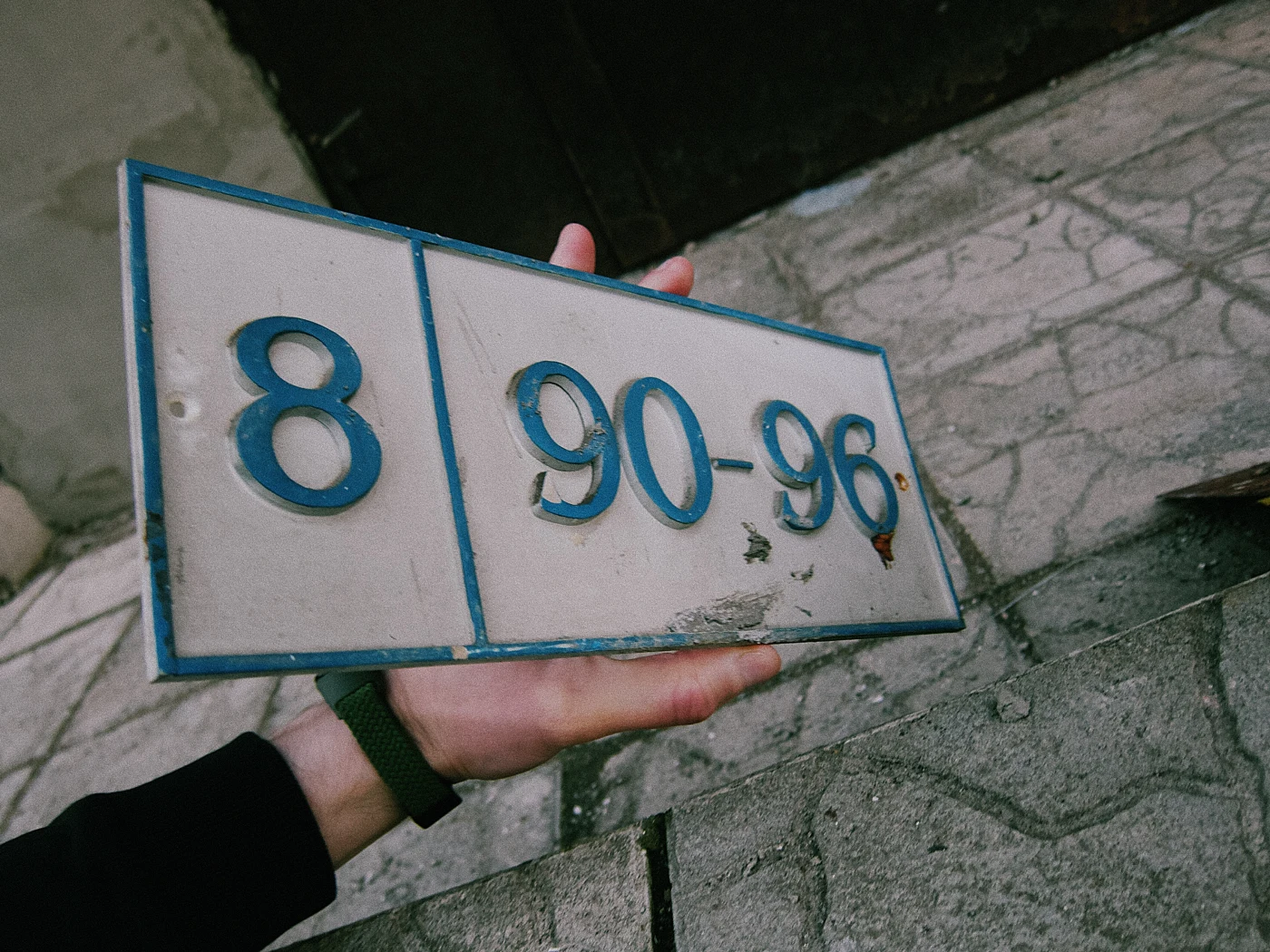 House number 8, 90–96, on an old sign held in a hand against a concrete surface.