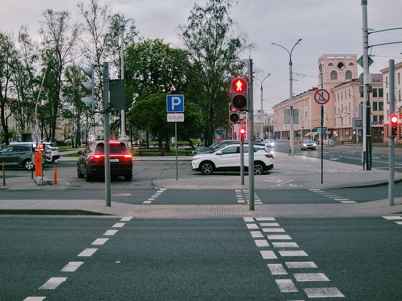 A traffic light with a red signal, cars at an intersection, a parking lot and a pedestrian crossing. Gray sky.